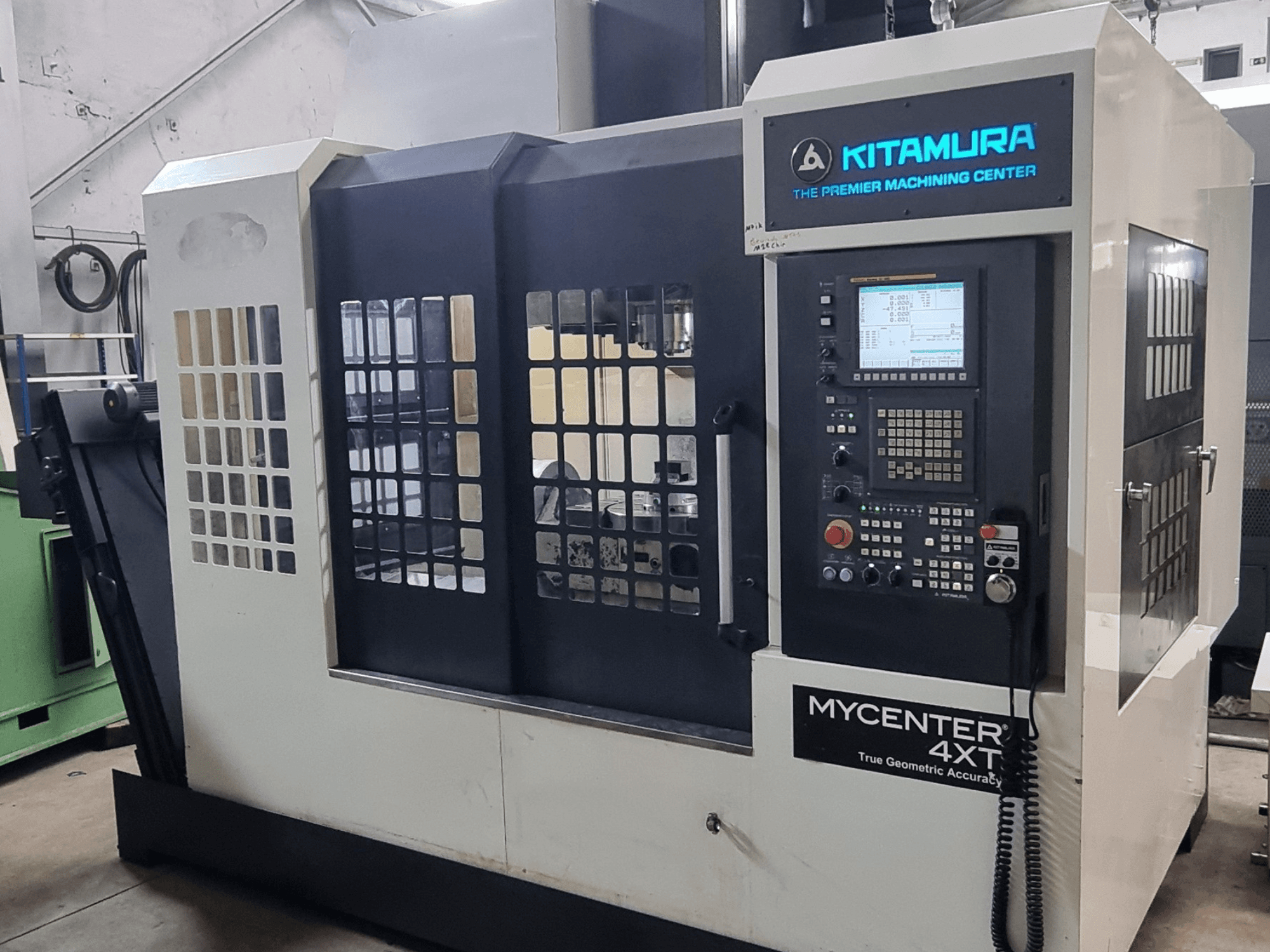 Alternatyvusis tekstas: „Kitamura MYCENTER 4XT“ vertikalusis apdirbimo centras, vaizdas iš priekio, kuriame matomas valdymo pultas ir staklių detalės dirbtuvės aplinkoje.