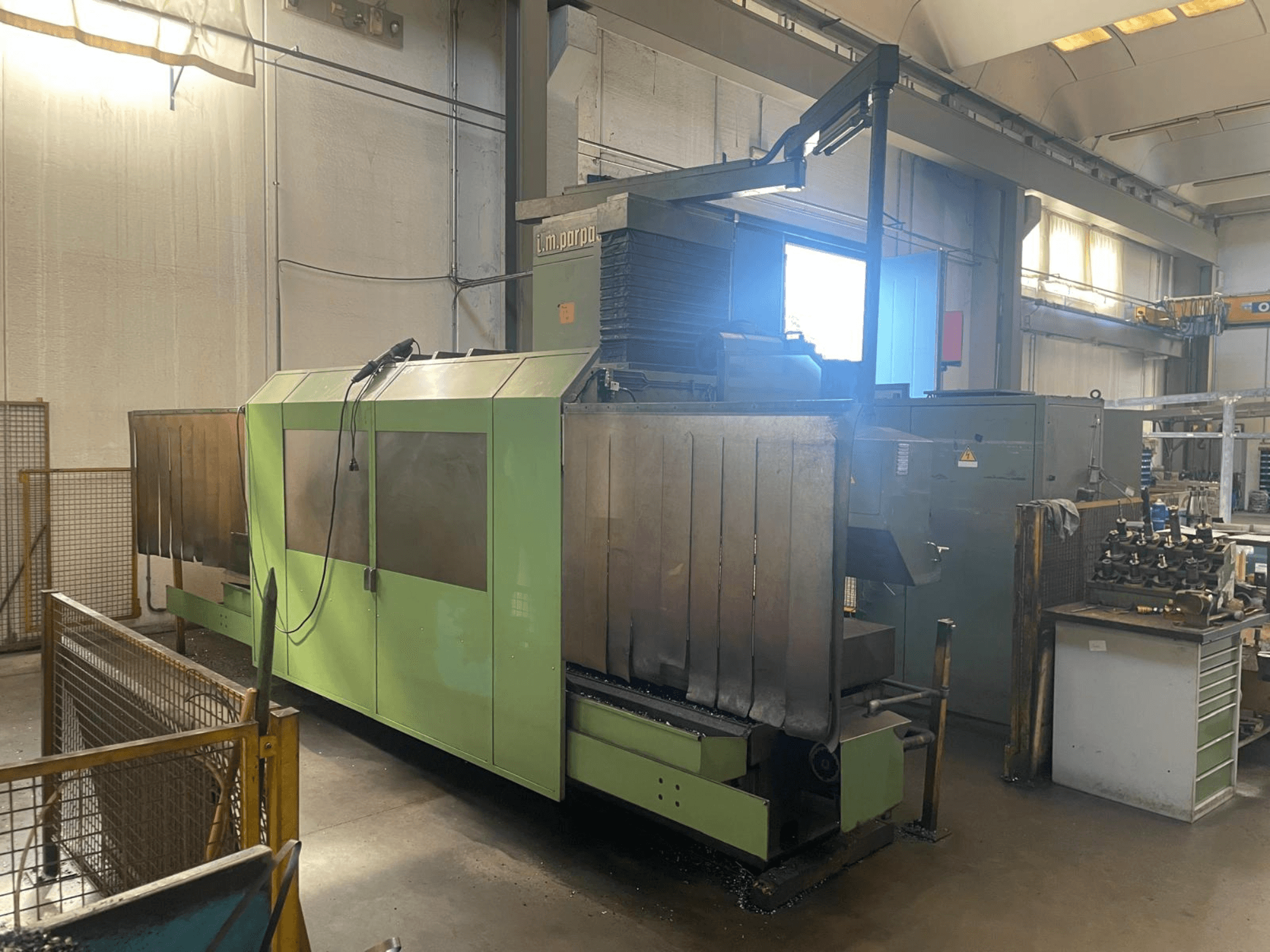 Žalios spalvos pramoninė mašina su matomais metaliniais komponentais, vaizdas iš priekio; šalia yra valdymo skydelis, saugos įspėjimai ir darbo vieta.