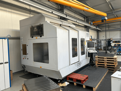 CNC staklės "Spinner VC 1650 CNC", vaizdas iš priekio, su valdymo skydeliu, dideliais langais ir šalia sukrautomis paletėmis dirbtuvėse.