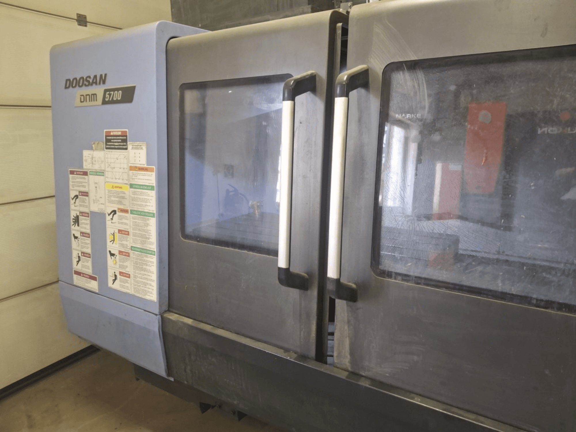 Vertikalaus apdirbimo centro DOOSAN DNM 5700 vaizdas iš priekio, matomos dvejos durys ir valdymo skydeliai su valdymo instrukcijomis.