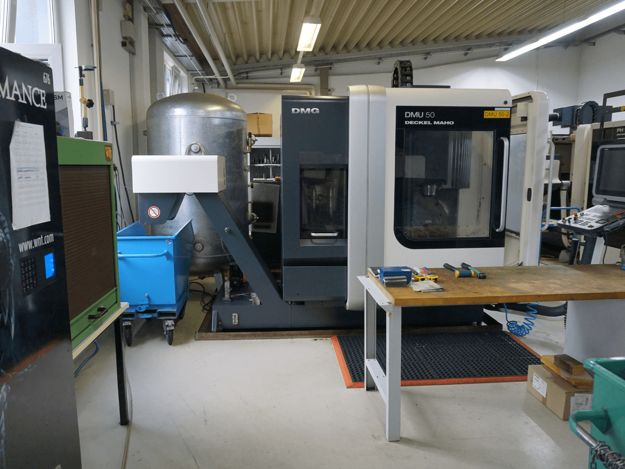 DMG DMU 50 frezavimo staklės dirbtuvėse, vaizdas iš priekio, su įrankiais ant medinio stalo ir šalia esančia mėlyna šiukšliadėže ant ratukų.