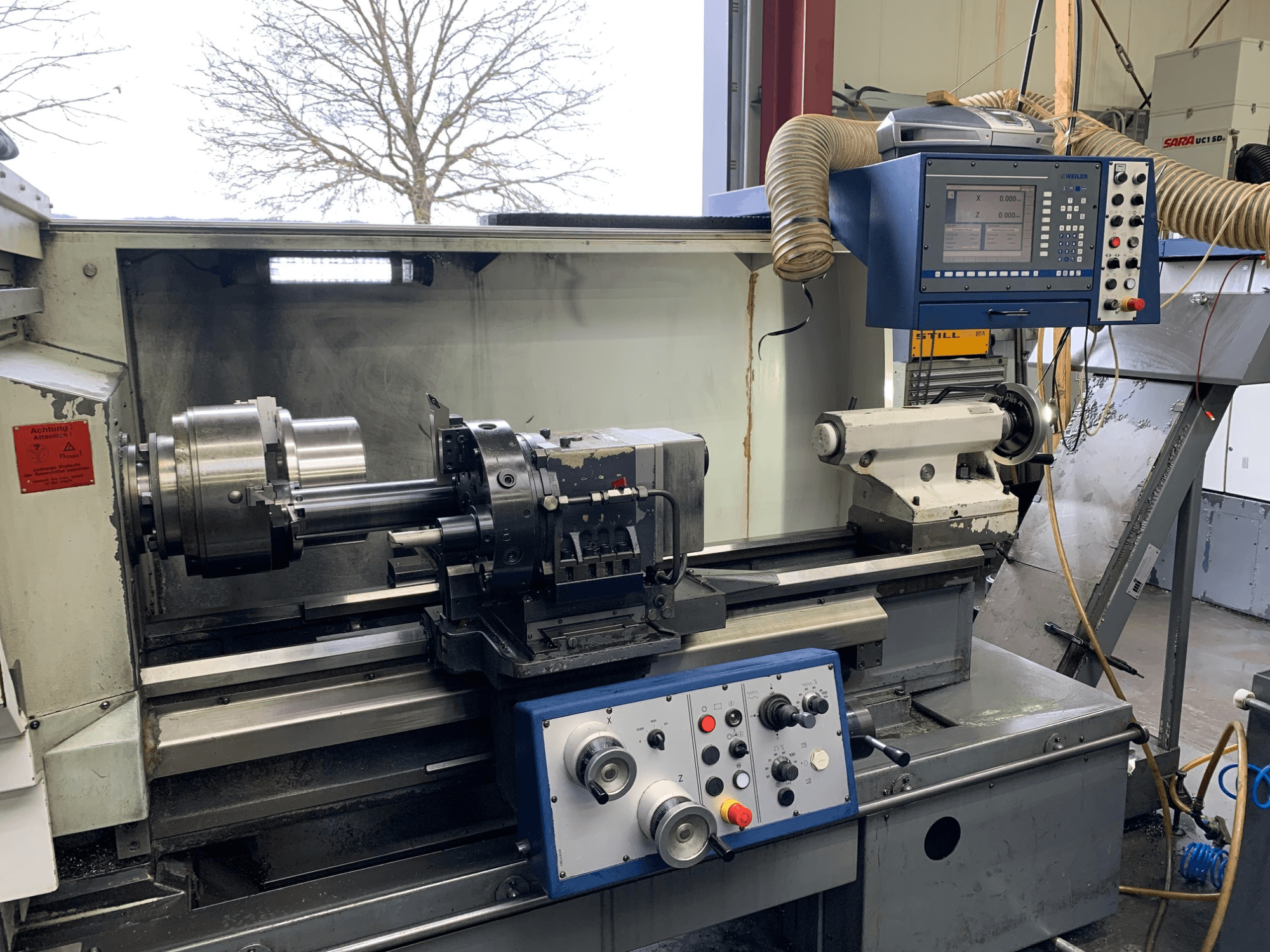 Weiler E55X1000 horizontalios tekinimo staklės dirbtuvėse, kuriose matomas valdymo pultas, tekinimo staklės ir įrankių laikiklis. Vaizdas iš priekio.