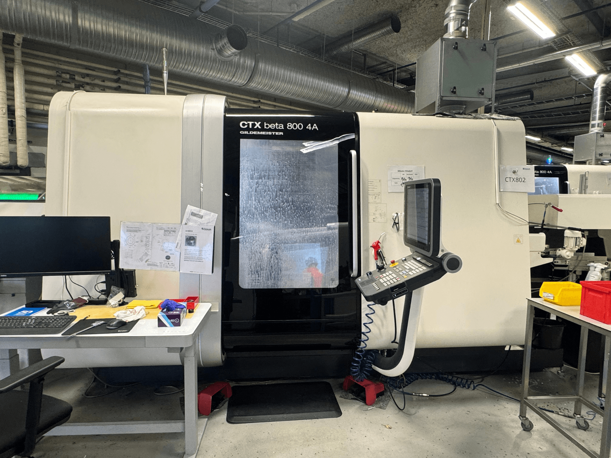 CTX beta 800 4A CNC staklės dirbtuvėse, šoninis vaizdas, kuriame matomas valdymo skydelis ir įrankiais bei dokumentais užgriozdinta darbo vieta.