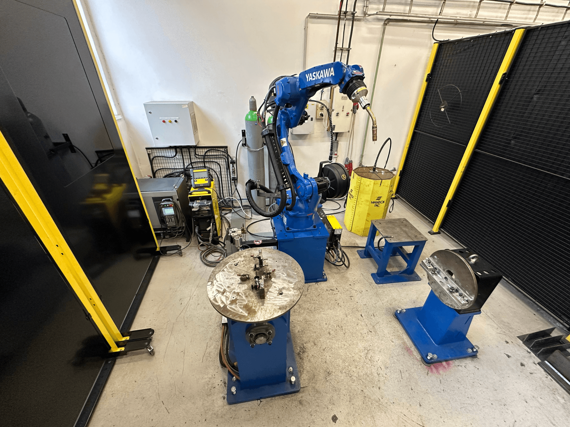Mėlynos spalvos "Yaskawa" roboto ranka su dviem darbo stalais, suvirinimo įranga ir apsauginiais barjerais; pramoninės darbo vietos vaizdas iš priekio.