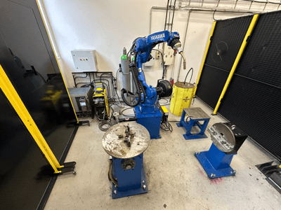 Mėlynos spalvos "Yaskawa" roboto ranka su dviem darbo stalais, suvirinimo įranga ir apsauginiais barjerais; pramoninės darbo vietos vaizdas iš priekio.