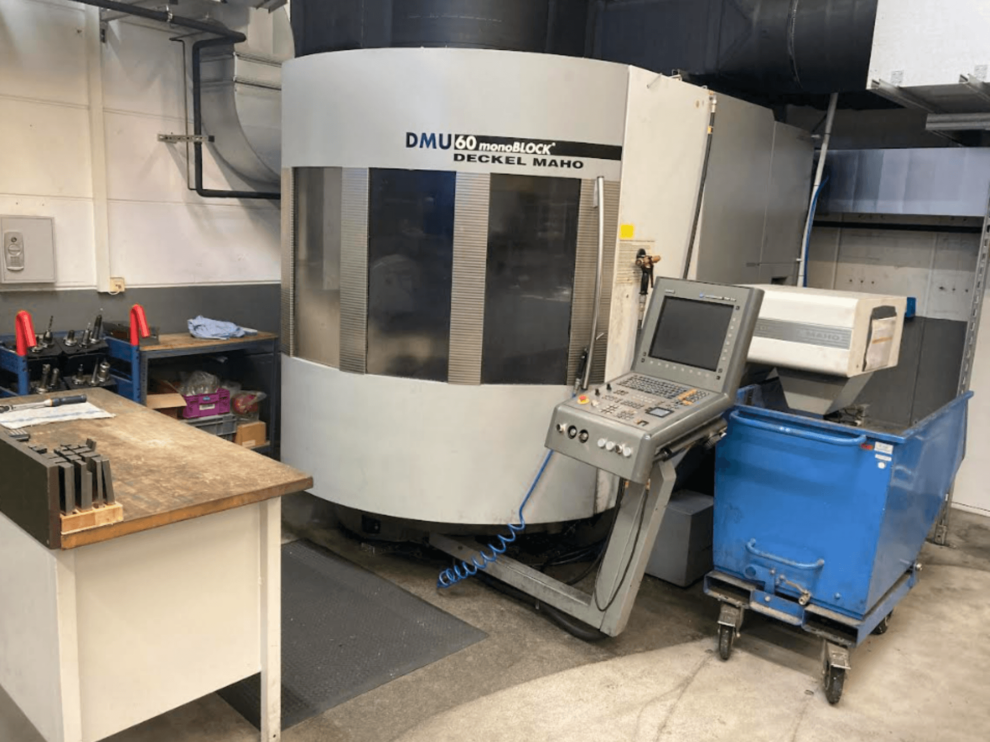 DMG MORI DMU 60 Monoblokinis vertikalusis apdirbimo centras, vaizdas iš priekio, matomas valdymo skydelis, darbo vieta ir mėlynas įrankių vežimėlis.