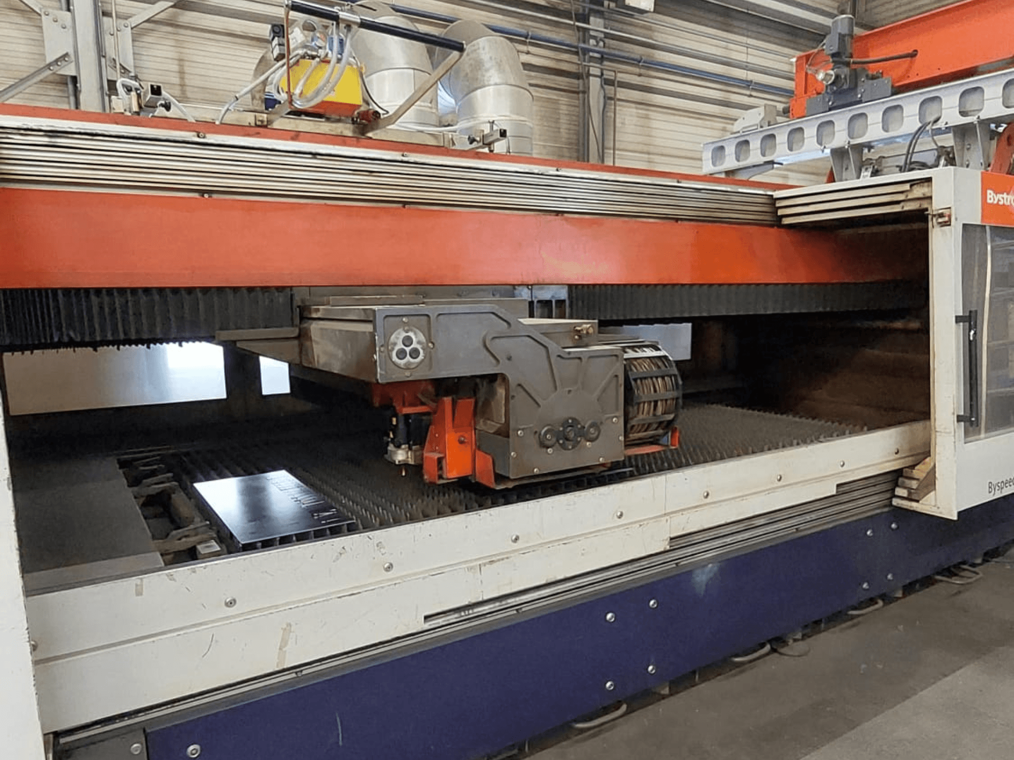 „Bystronic BySpeed 3015“ CO₂ lazerio pjovimo staklės, vaizdas iš priekio, kuriame matoma pjovimo galvutė ir metalinis lakštas ant pjovimo stalo.