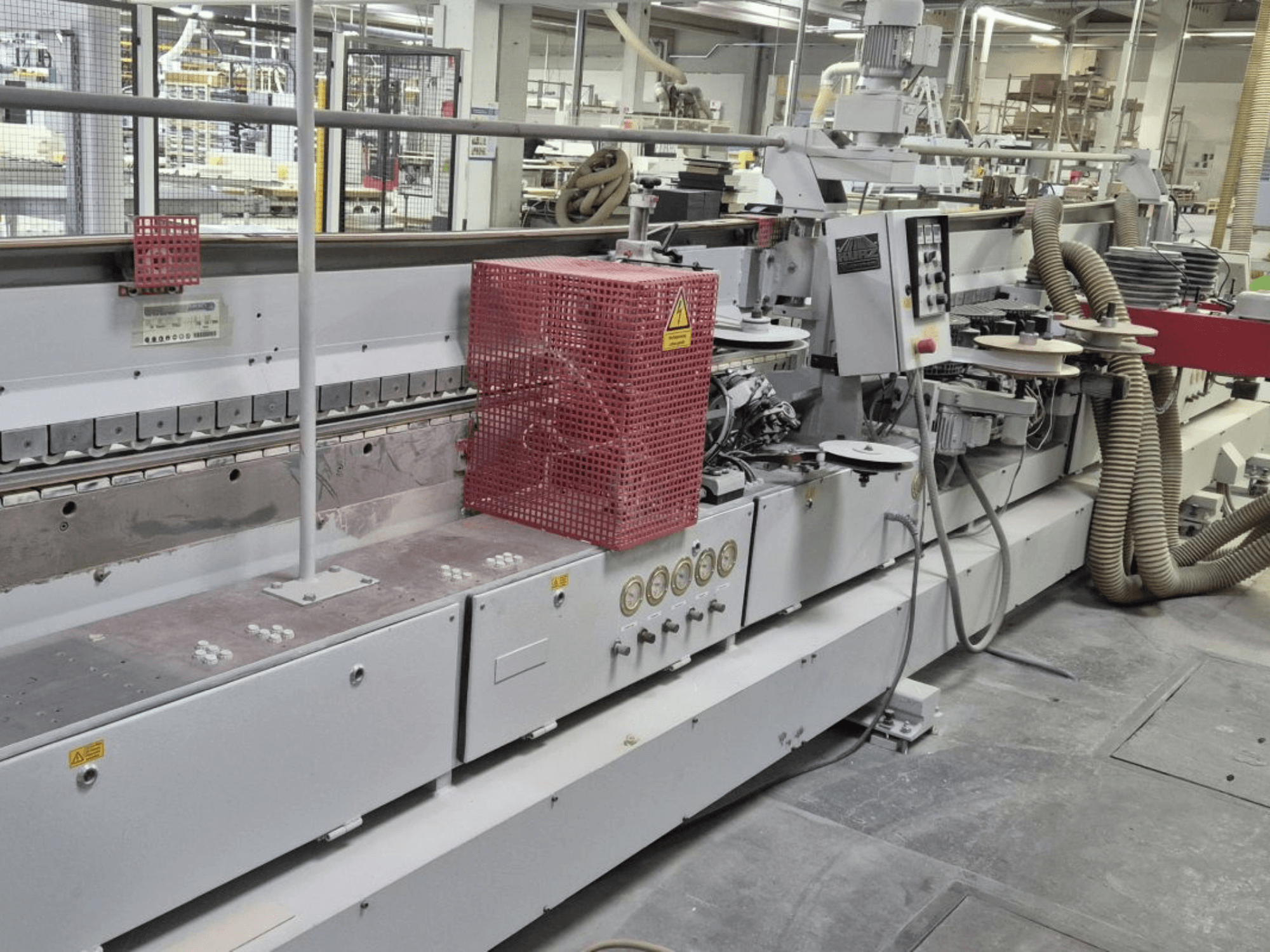 Kraštų juostų klijavimo mašina su valdymo pultu, siurbimo žarnomis ir raudonu apsauginiu dangčiu, žiūrint iš šono.