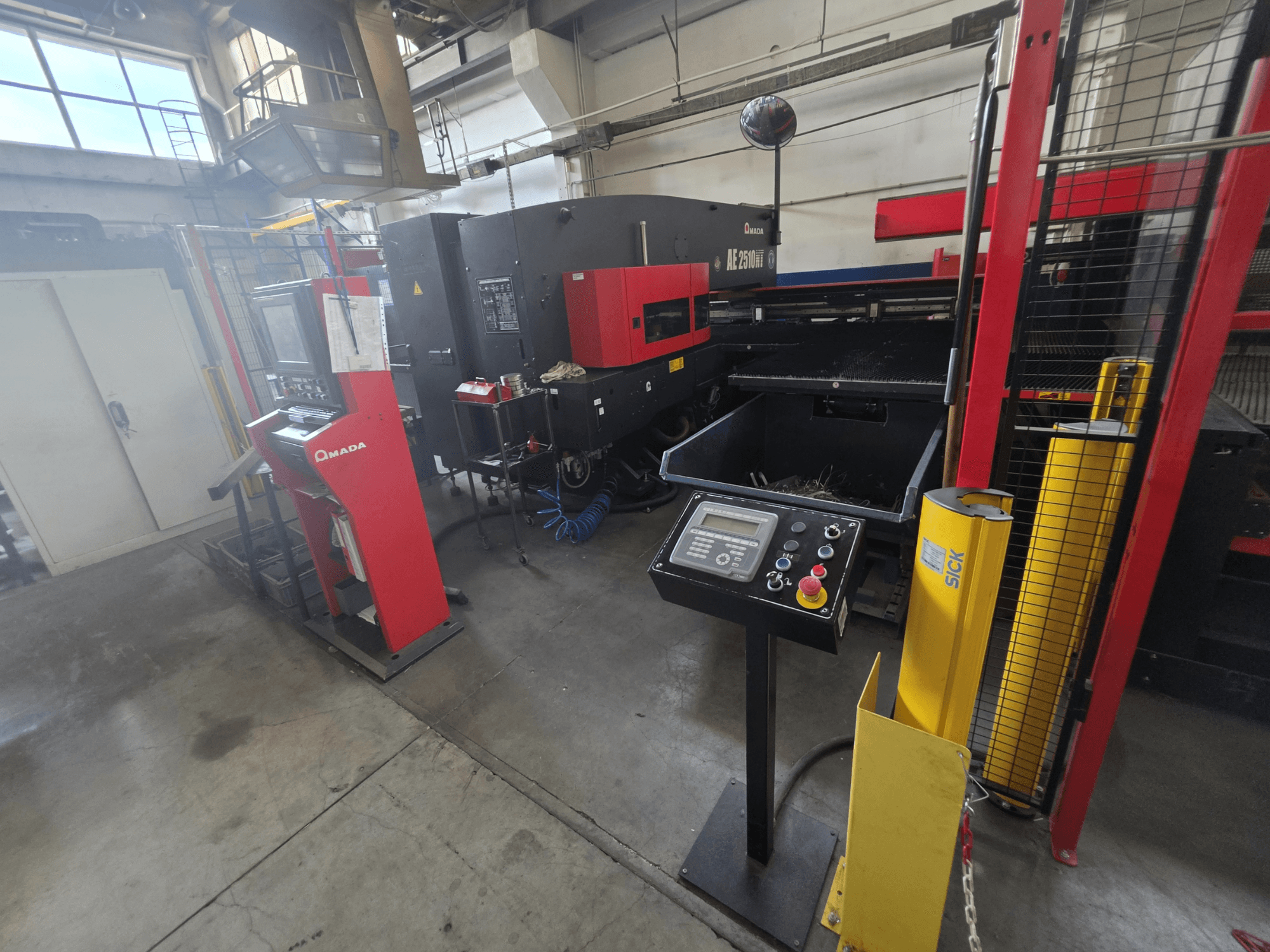 CNC štampavimo staklės „AMADA AE-2510NT“, vaizdas iš priekio su valdymo pultu, saugos įranga ir aplinkinėmis pramoninėmis patalpomis.