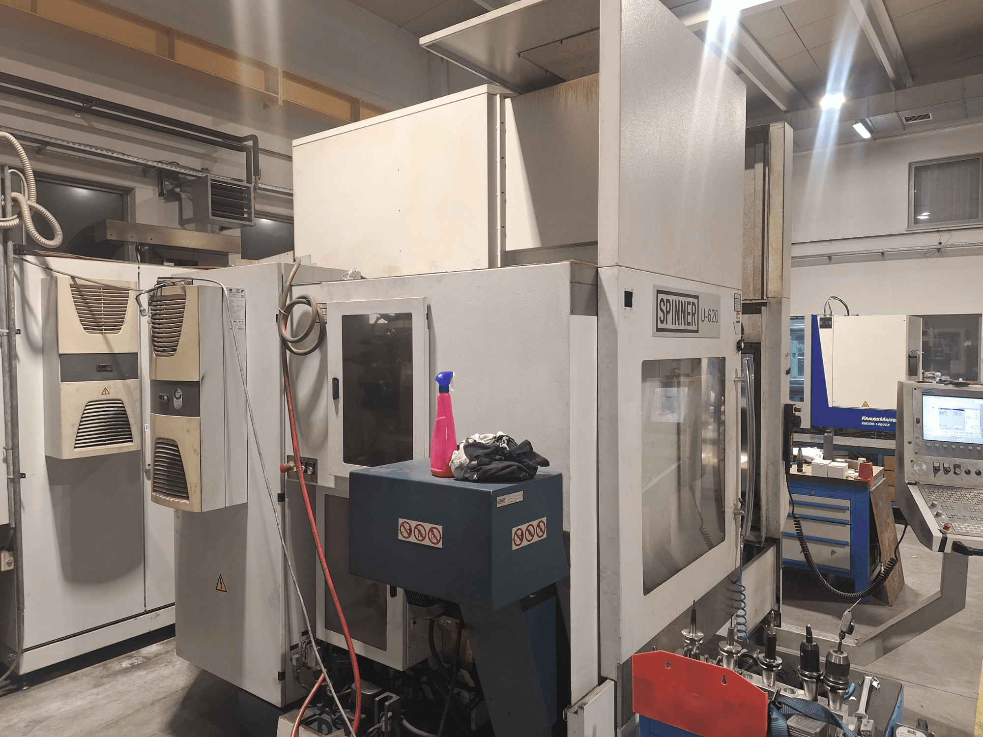 CNC staklės "Spinner U-620", parodytos dirbtuvėse iš priekio, su valdymo pultu ir įvairiais įrankiais ant šalia esančio darbastalio.