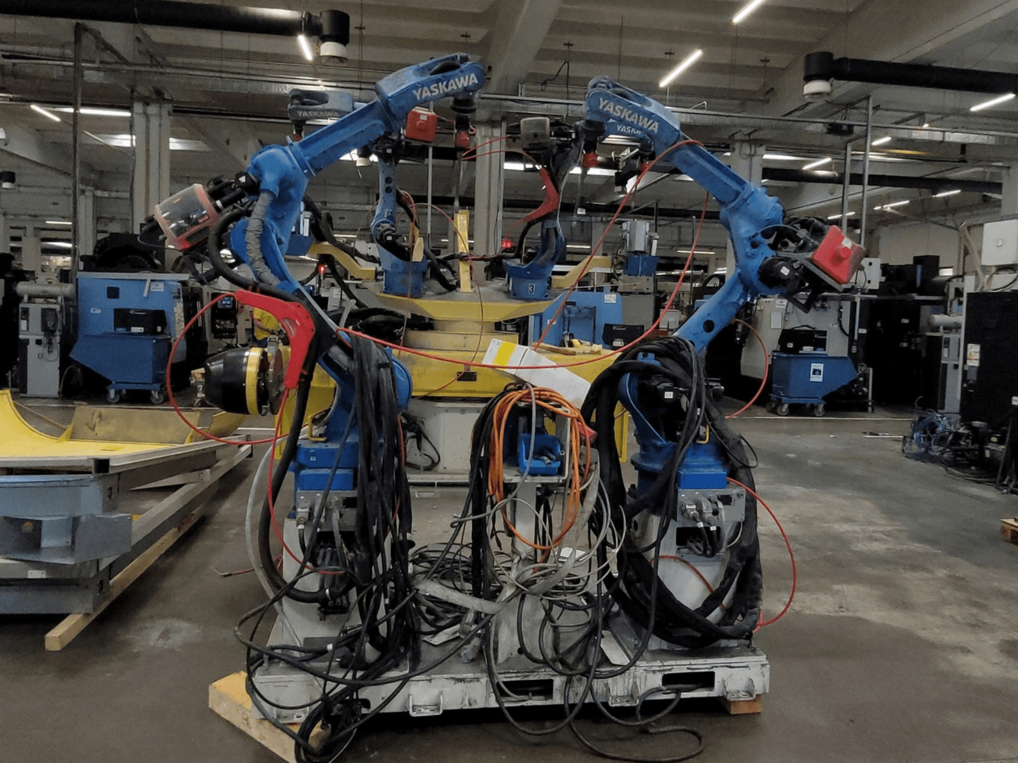 „Yaskawa“ robotizuota suvirinimo linija RWC L1_OS 15330, vaizdas iš priekio, su dviem mėlynais robotais, daugybe kabelių ir suvirinimo įrankiais.