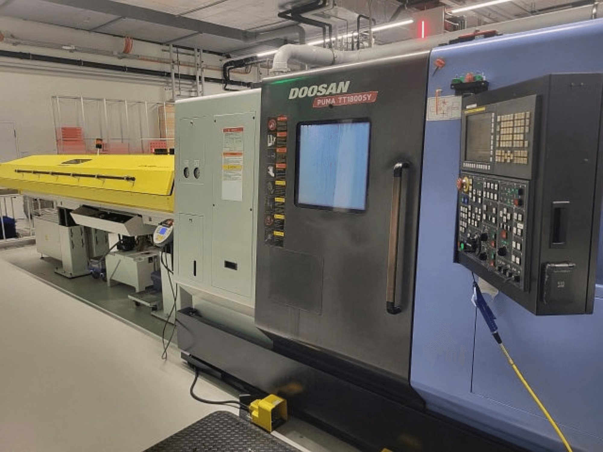 „DOOSAN PUMA TT 1800 SY“ tekinimo ir frezavimo centras, vaizdas iš priekio su valdymo pultu ir geltonu dangčiu, pramoninė aplinka.