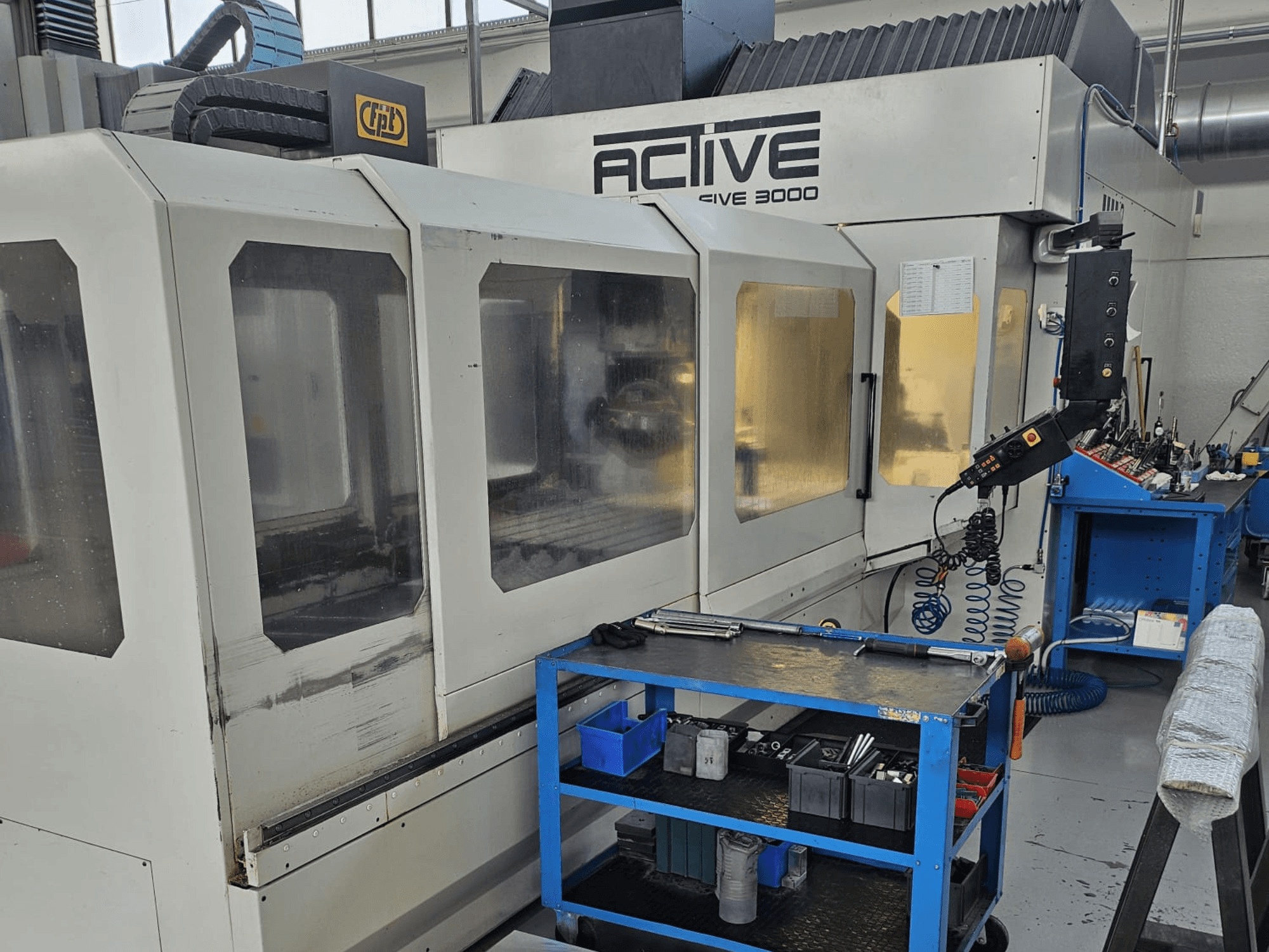 Aktyvios 3000 CNC staklės dirbtuvėse, vaizdas iš priekio su įrankiais ant mėlyno vežimėlio, viduje matomi valdikliai ir darbo zona.