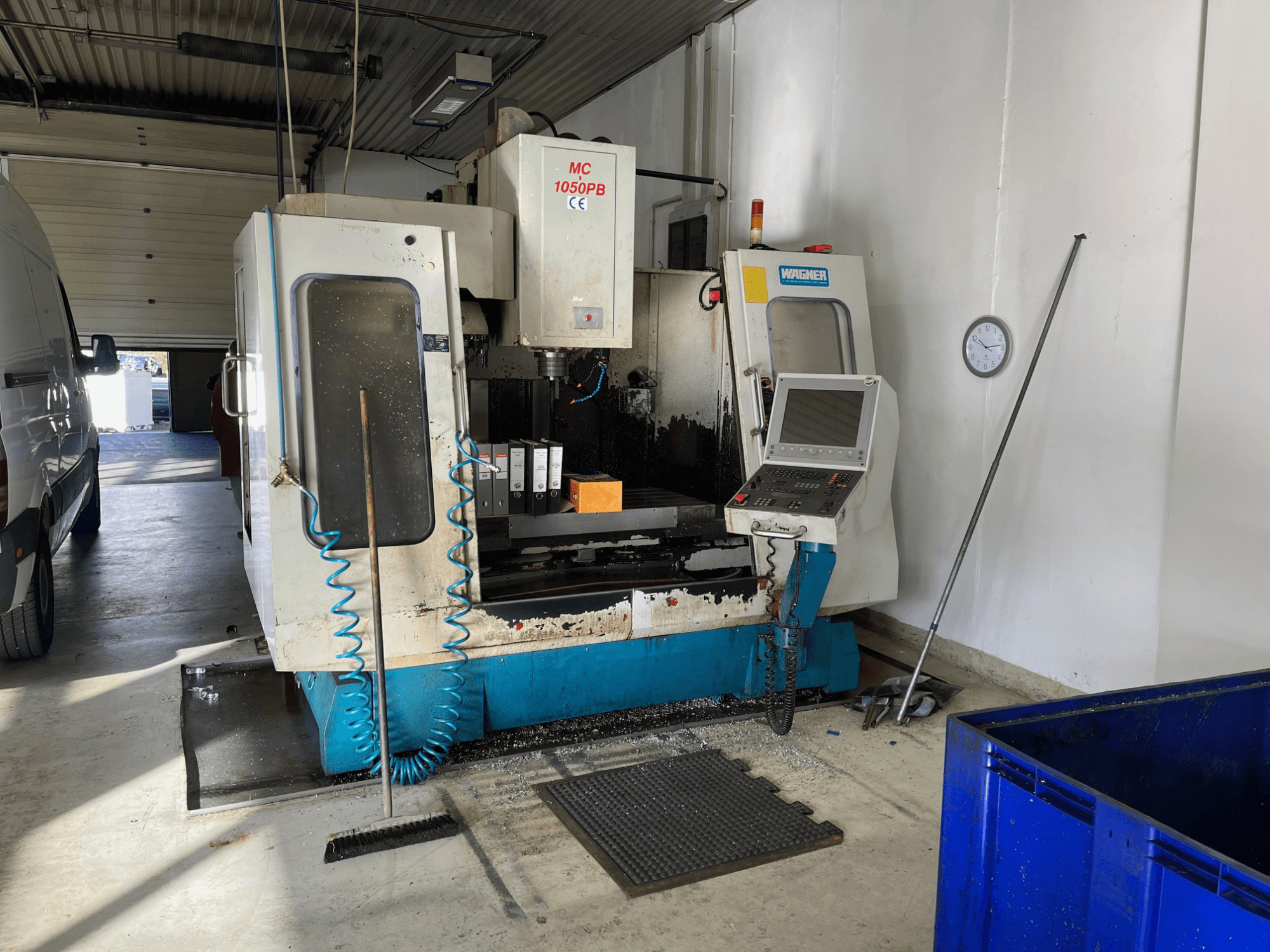 WAGNER MC 1050PB CNC staklės dirbtuvėse, vaizdas iš priekio, su valdymo pultu ir darbo zona; matoma mėlyna oro tiekimo žarna ir valymo įrankiai.