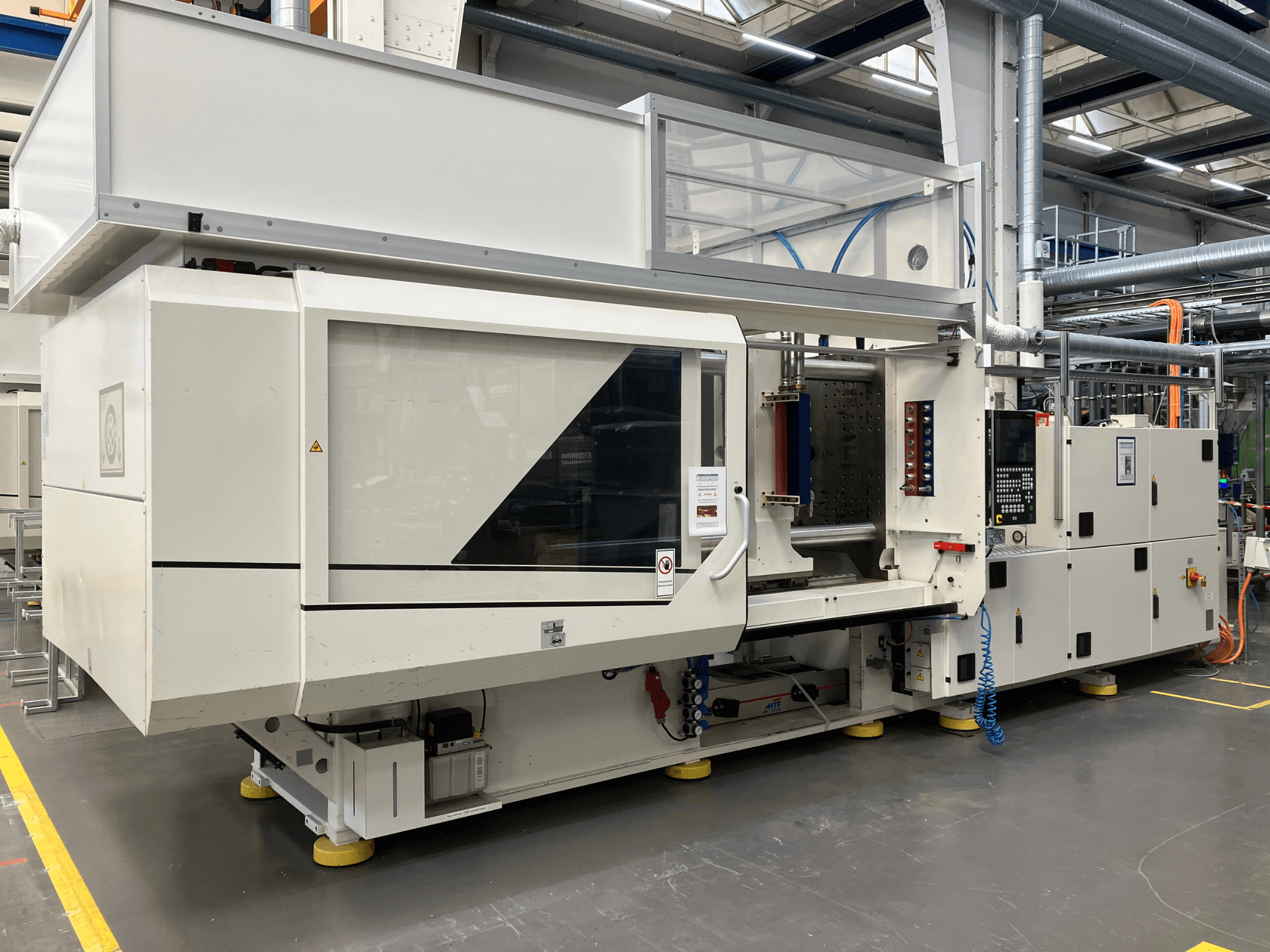 Šoninis AET modelio liejimo mašinos su valdymo skydeliu ir skaidriais dangteliais vaizdas pramoninėje aplinkoje.