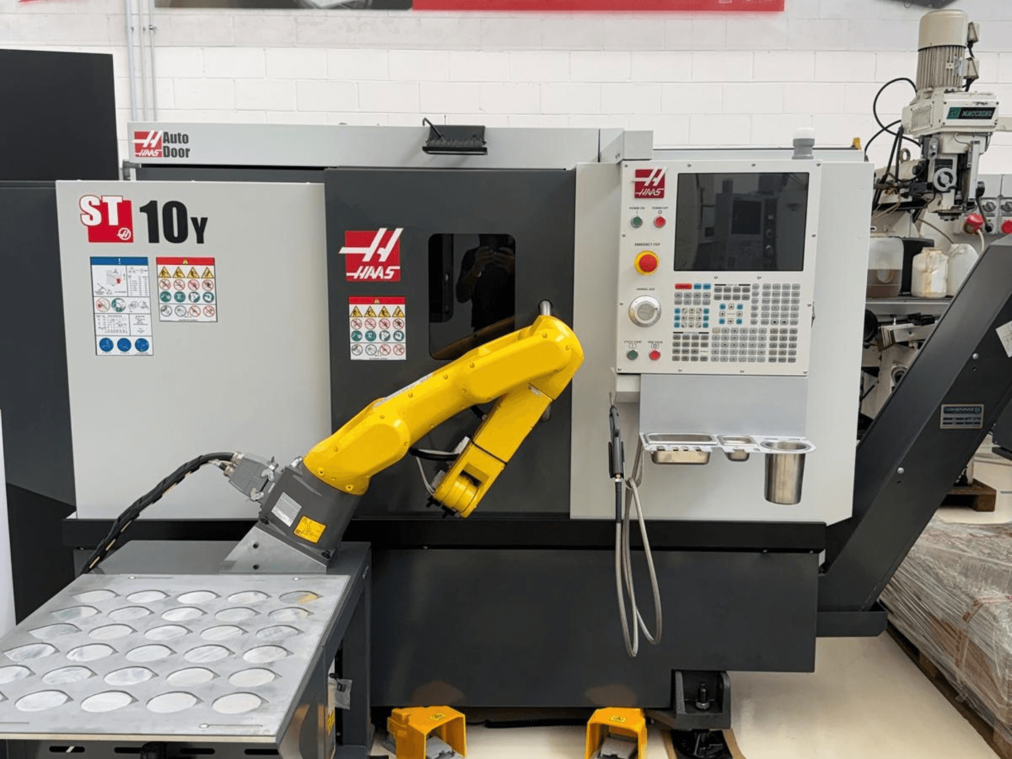 „Haas ST-10Y“ horizontalusis tekinimo staklės, matomos iš priekio, su robotine ranka ir valdymo pultu su įrankių laikiklių dėžėmis.