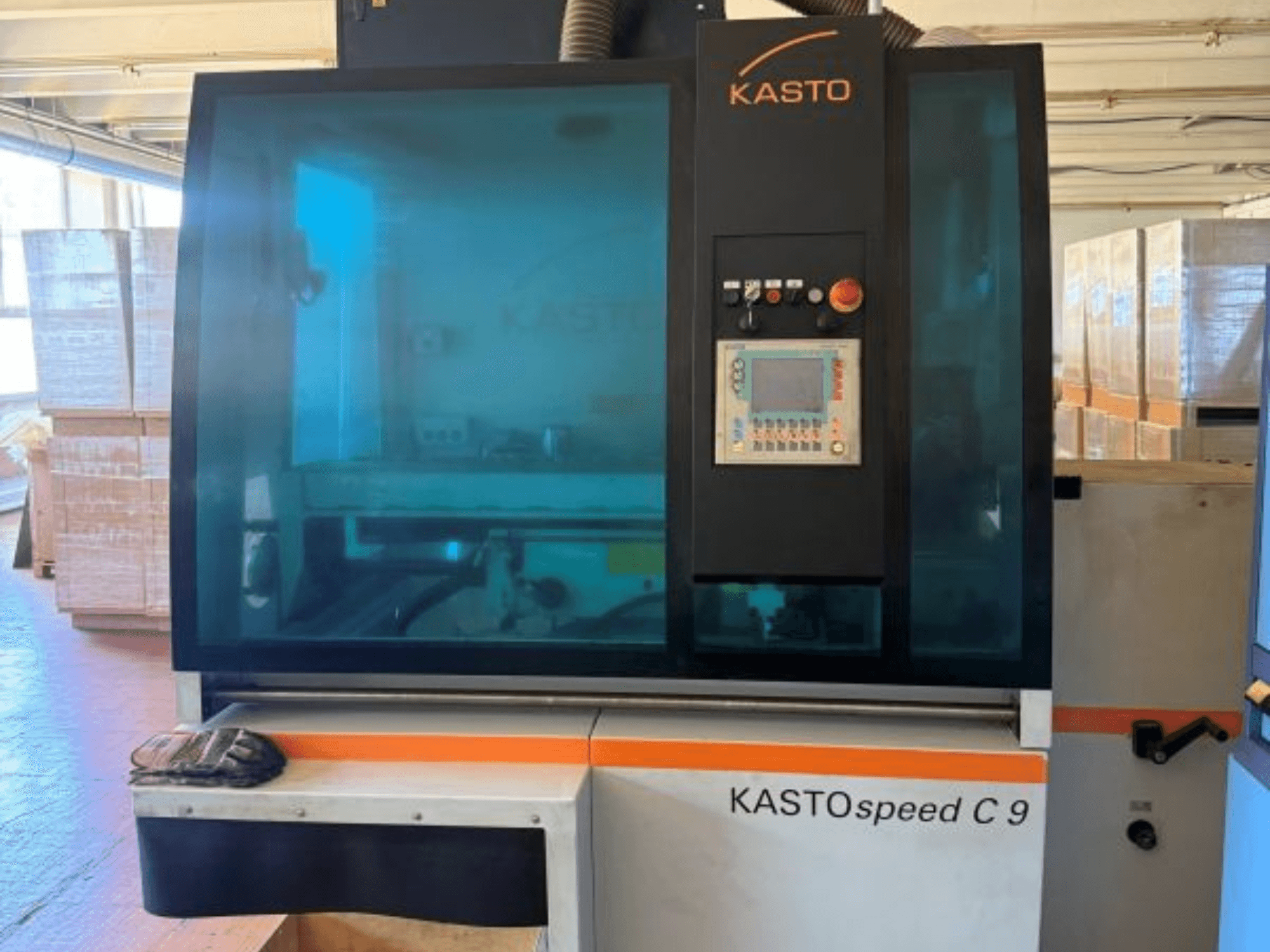 KASTO KASTOspeed C 9 apskrito pjūklo vaizdas iš priekio, kuriame matomas valdymo skydelis ir apsauginis stiklas, o fone – medinės dėžės.