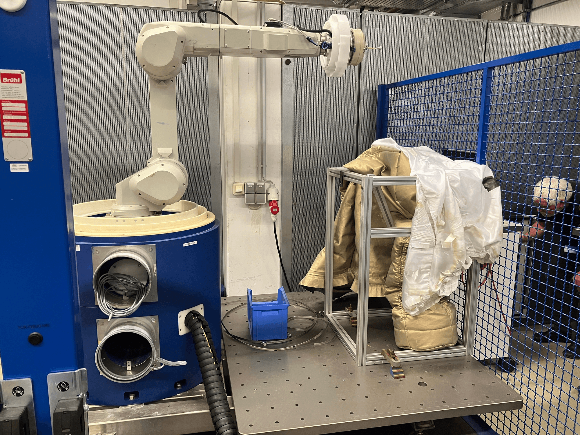 Roboto ranka prie mėlynos kameros, laikanti baltos ir auksinės spalvos kostiumą ant metalinio rėmo, vaizdas iš šono su apsauginio tinklelio fonu.