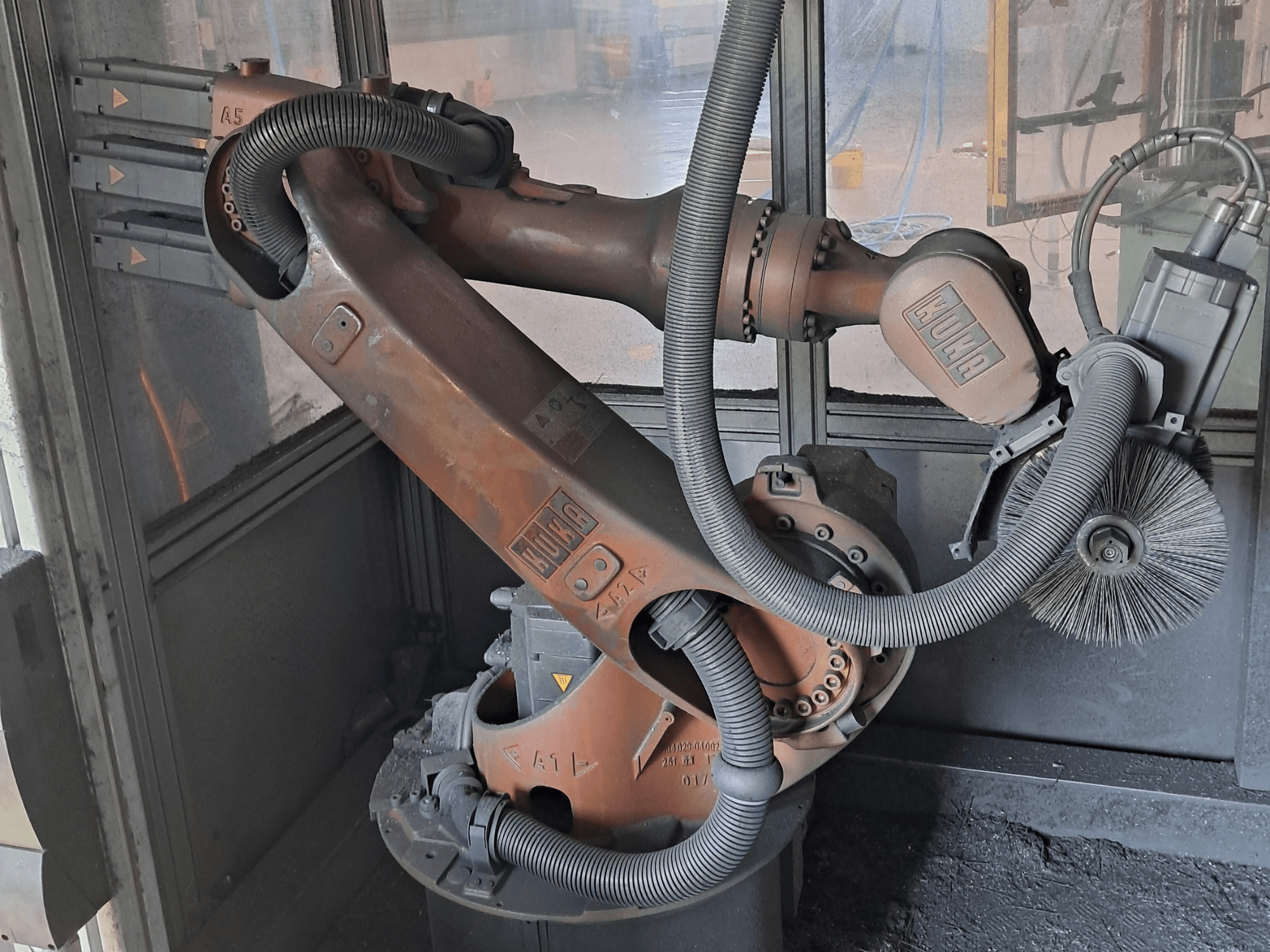„KUKA KR 16-2“ robotinė ranka, vaizduojama iš šono, kurioje matomi kabeliai, šepetėliai ir mechaniniai jungiamieji elementai, įkomponuota darbo aplinkoje.