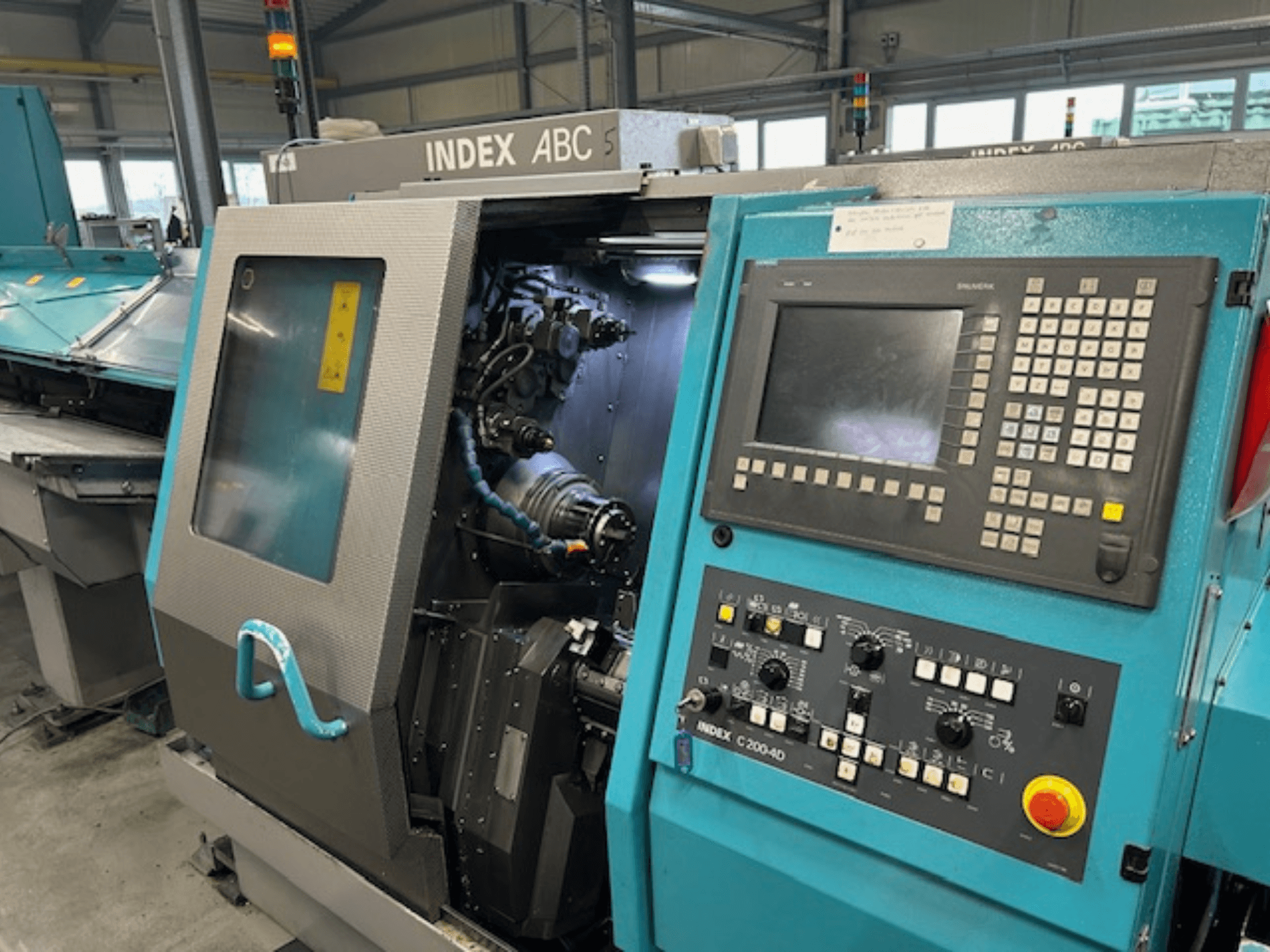 INDEX C200-AD CNC tekinimo staklių priekinis vaizdas su valdymo skydeliu, matomais vidiniais komponentais ir saugos elementais.