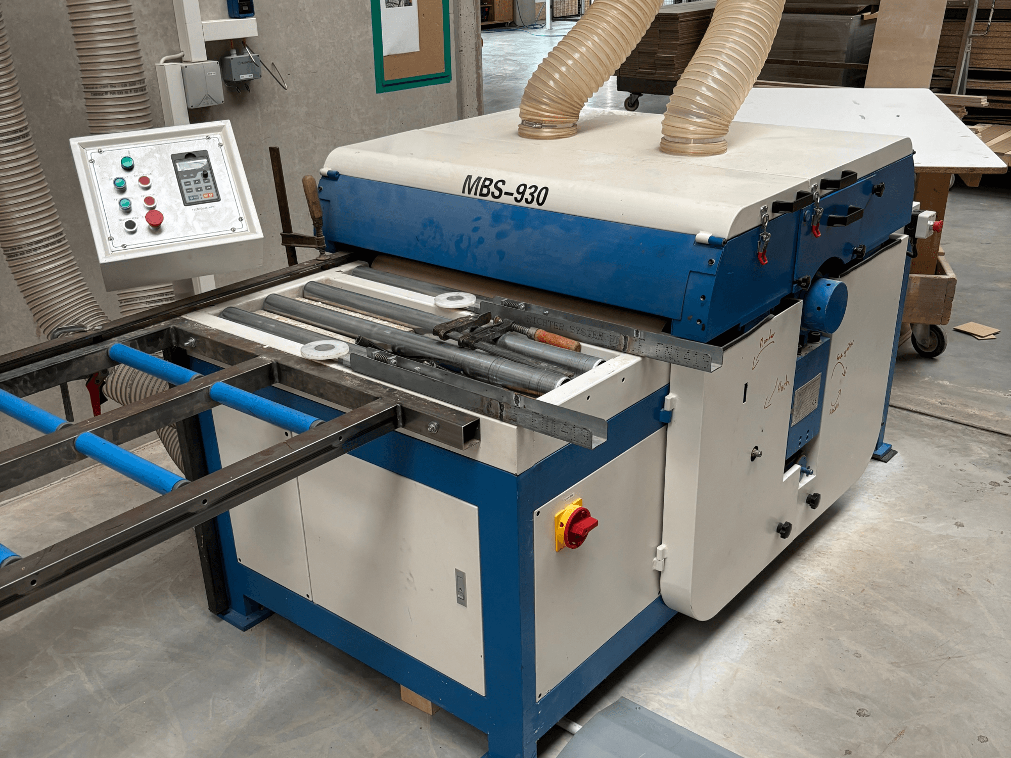 MBS-930 medžio apdirbimo staklės su valdymo pultu ir ritinėliais, žiūrint iš šono, su dulkių nusiurbimo žarnomis.