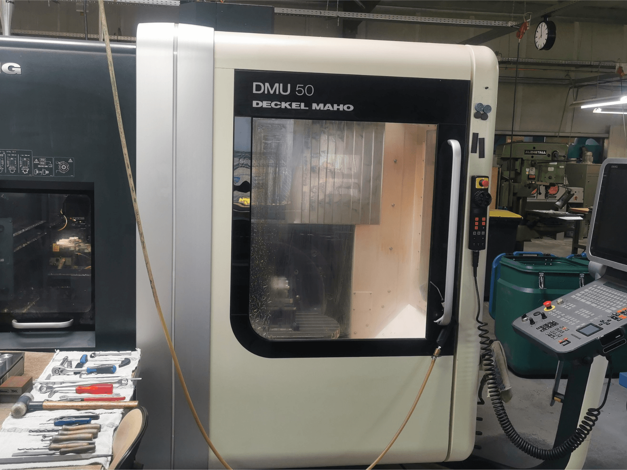 DMU 50 deckel maho CNC staklės, žiūrint iš priekio, su valdymo pultu ir permatomomis durelėmis. Įrankiai yra netoliese.