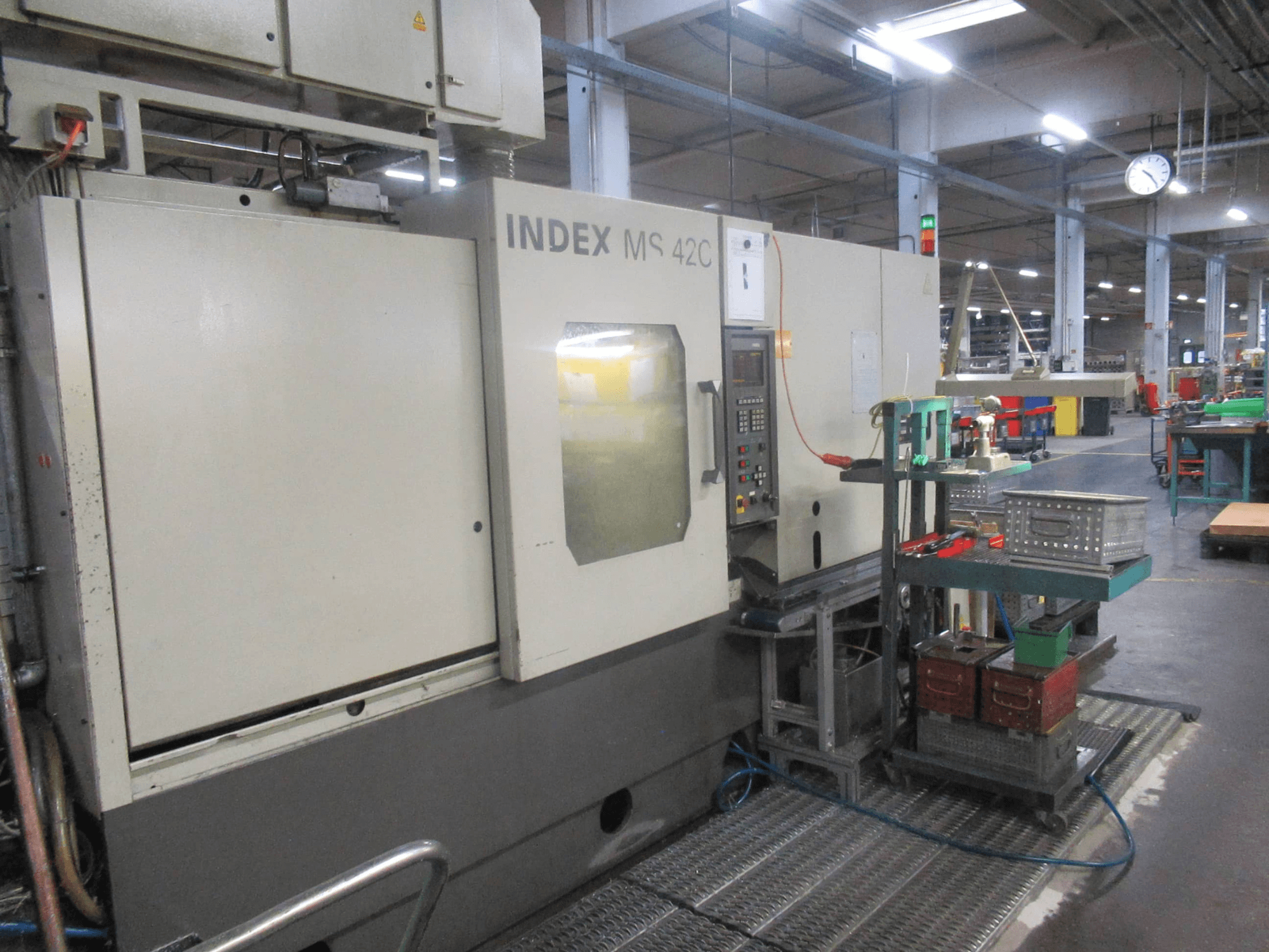 INDEX MS 42C CNC staklės dirbtuvėse, šoninis vaizdas, kuriame matomas valdymo skydelis ir įrankių laikymo vieta ant ratinio vežimėlio.