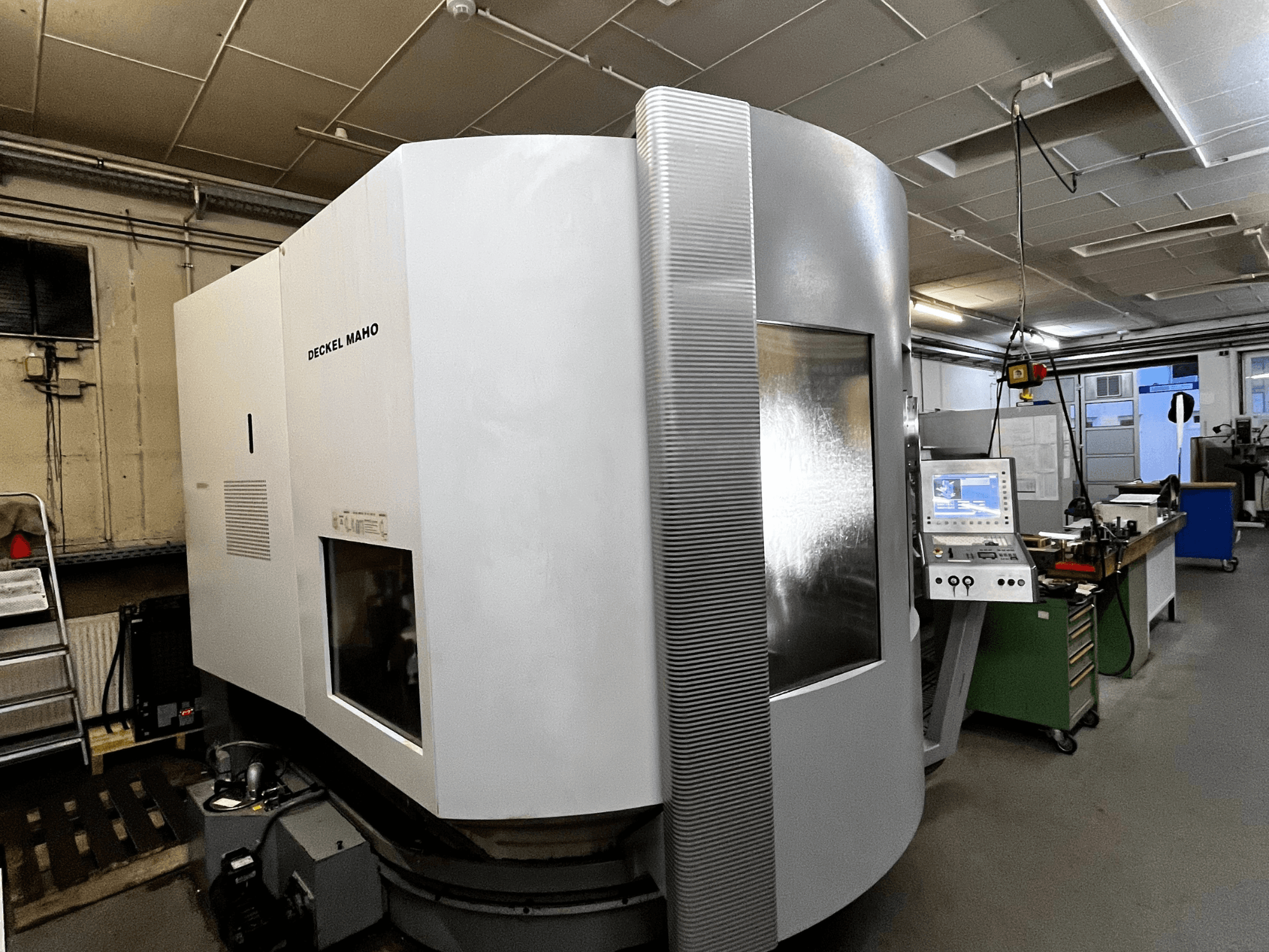 Deckel Maho CNC staklės, vaizdas iš priekio, balta išorė, matomas valdymo skydelis, dirbtuvių aplinka.