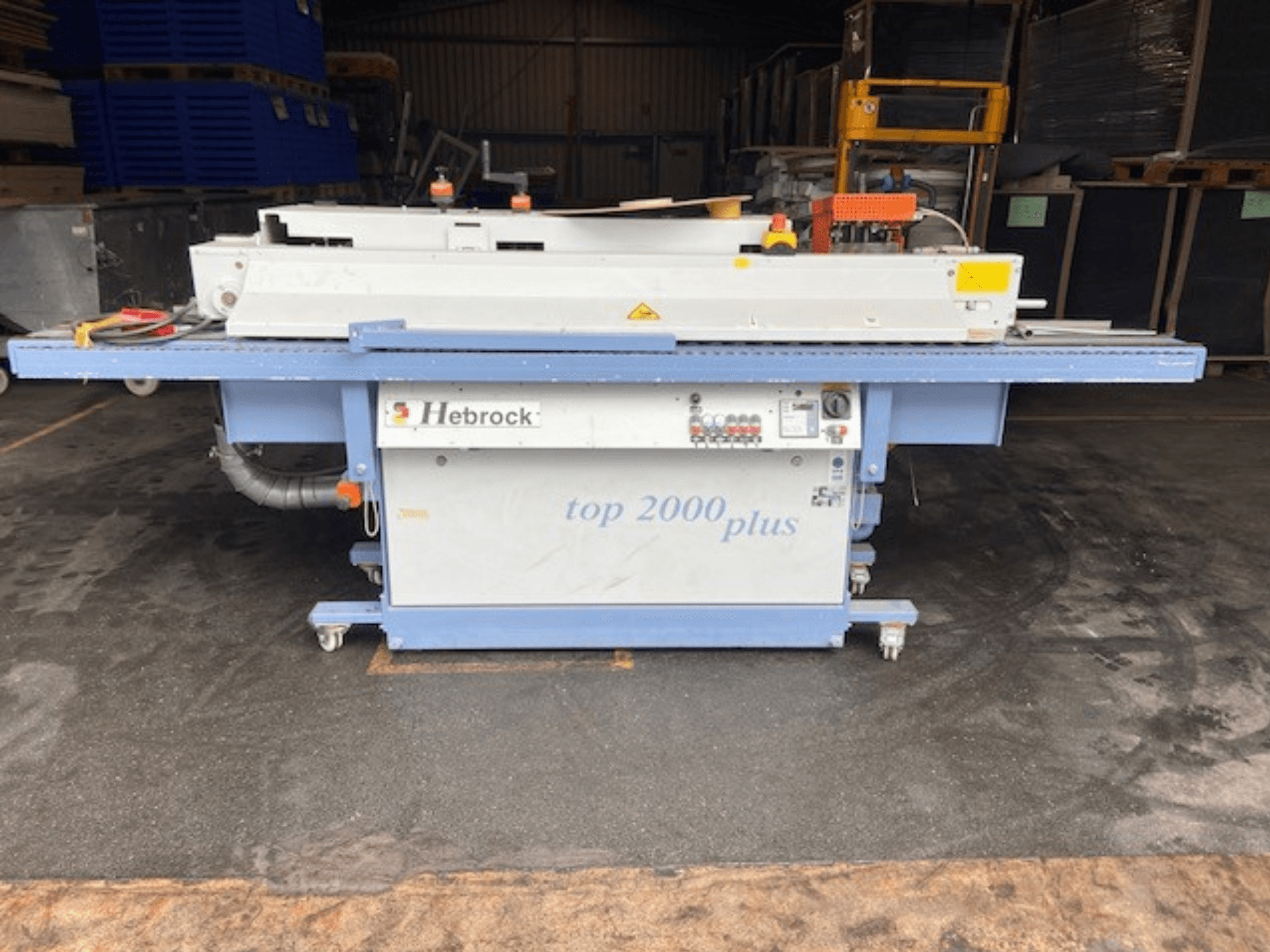 "Hebrock Top 2000 Plus", vaizdas iš priekio; pramoninė elektrinė mašina su valdymo skydeliu, judriais ratukais ir matomomis saugos etiketėmis.