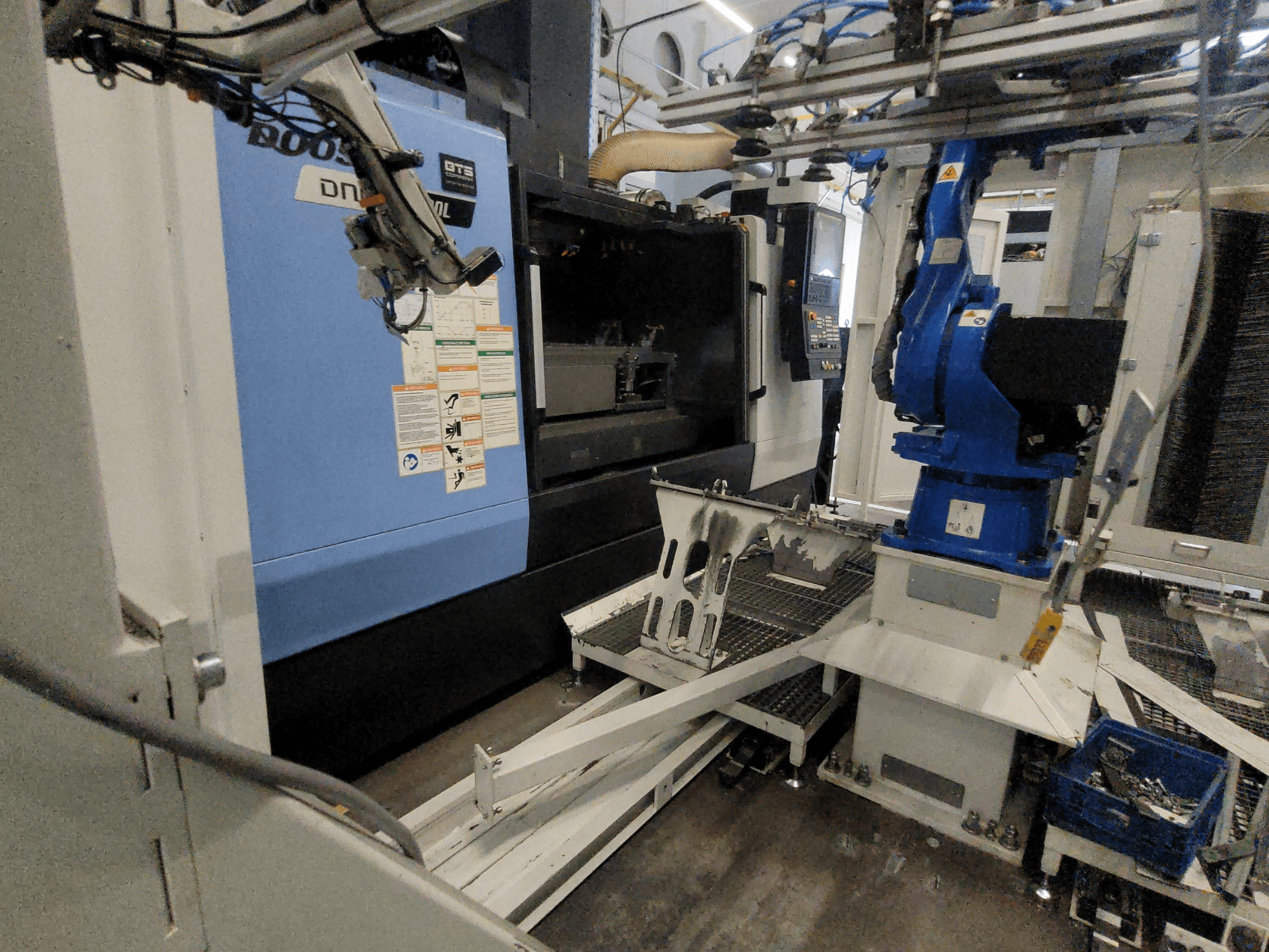 „DOOSAN DNM 6700L“ vertikaliojo apdirbimo centro vaizdas iš šono, kuriame matoma robotinė ranka ir valdymo pultas, atspindintis įrenginio konfigūraciją.