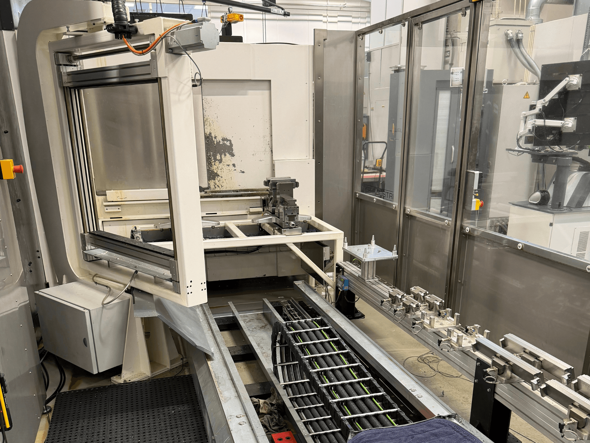 Automatizuota pramoninė darbo vieta, kurioje matoma roboto ranka, bėgių sistema ir valdymo pultas iš šono.