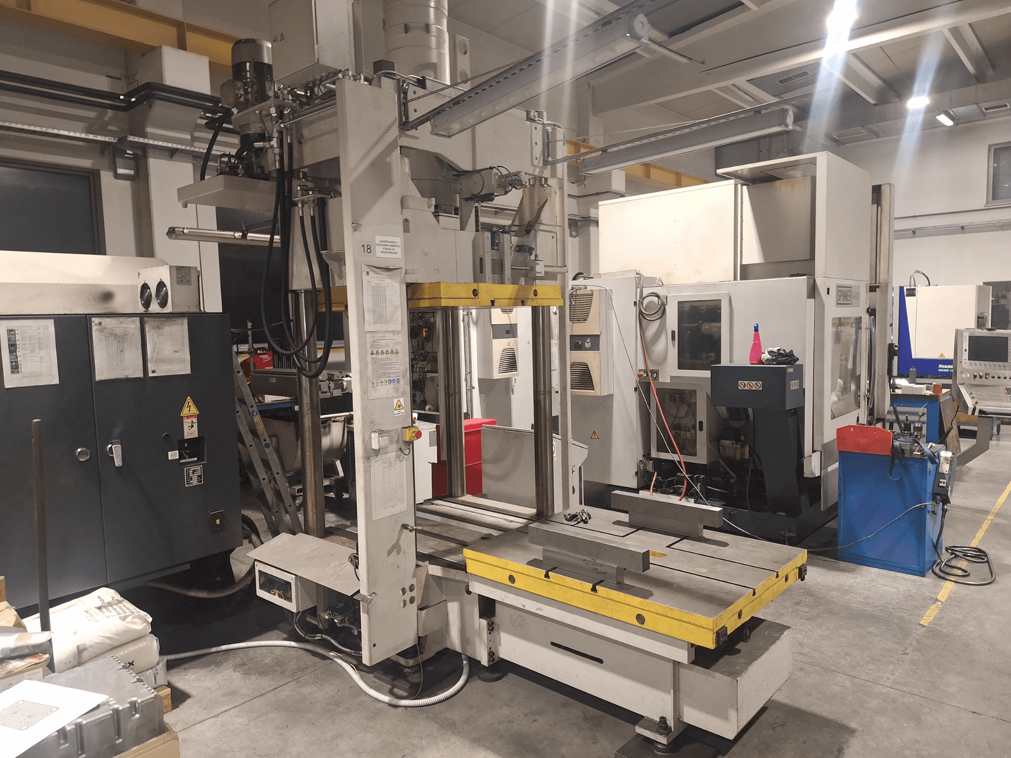 Horizontaliosios frezavimo staklės "Synergic", vaizdas iš priekio; platus stalas ir įvairūs valdymo skydeliai dirbtuvėse.