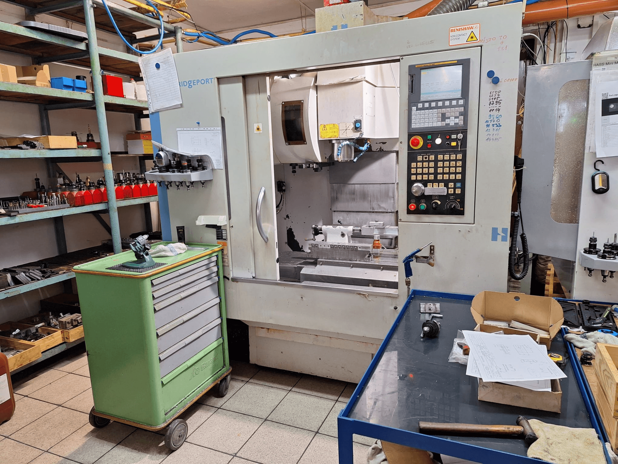 „Hardinge Bridgeport GX 710“ vertikaliojo apdirbimo centro vaizdas iš priekio su valdymo pultu, įrankiais ir lentynomis dirbtuvės aplinkoje.