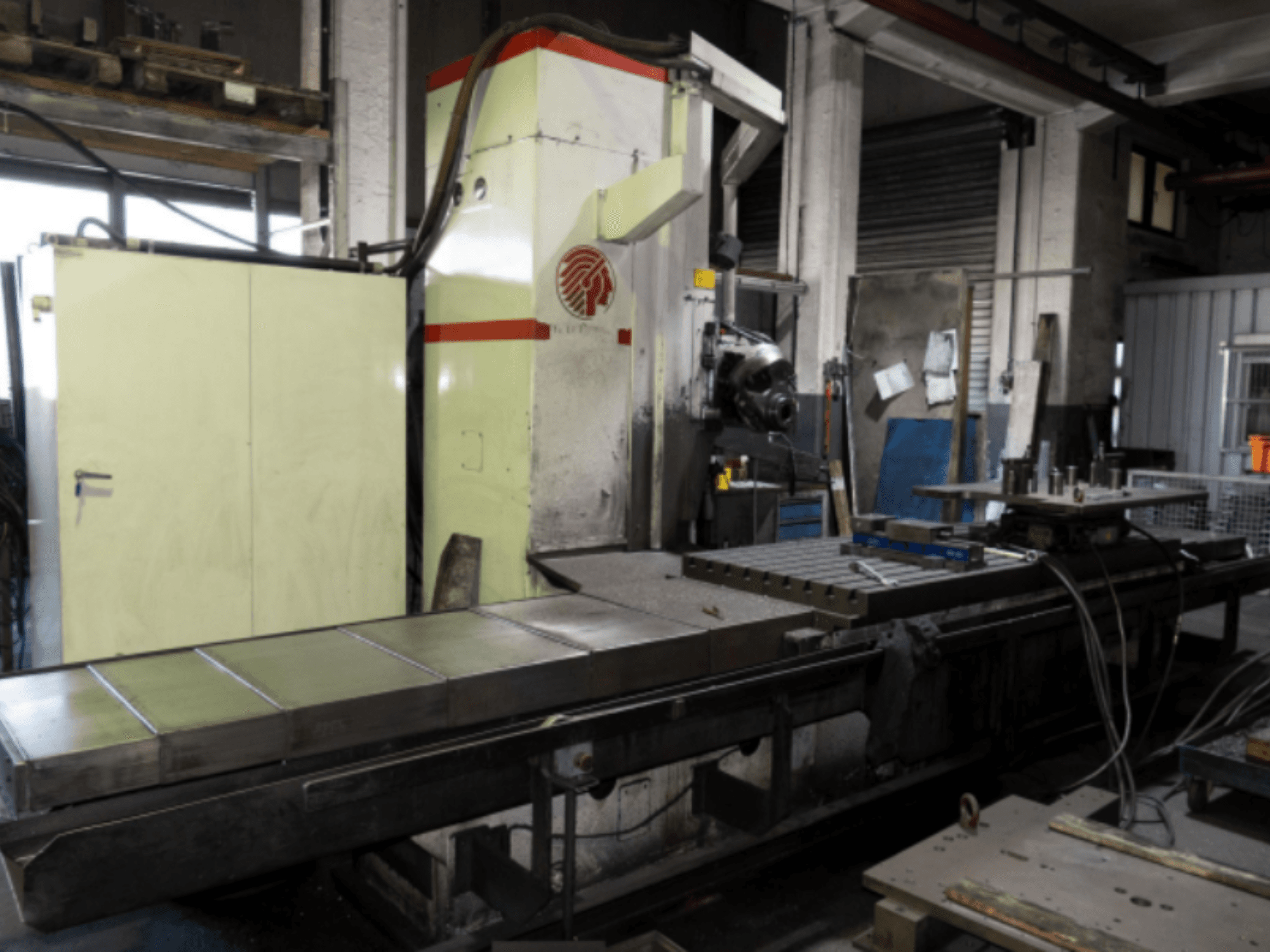 Pramoninės frezavimo staklės su dideliu stalu, šoninis vaizdas, demonstruojant valdymo įtaisus ir priedus dirbtuvėse.