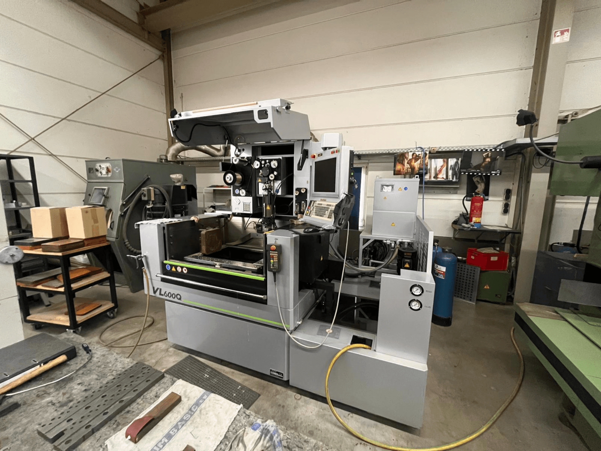 VL600Q CNC staklės dirbtuvėse, vaizdas iš priekio, su valdymo skydeliu, padėta medžiaga ir įvairiais įrankiais šalia.