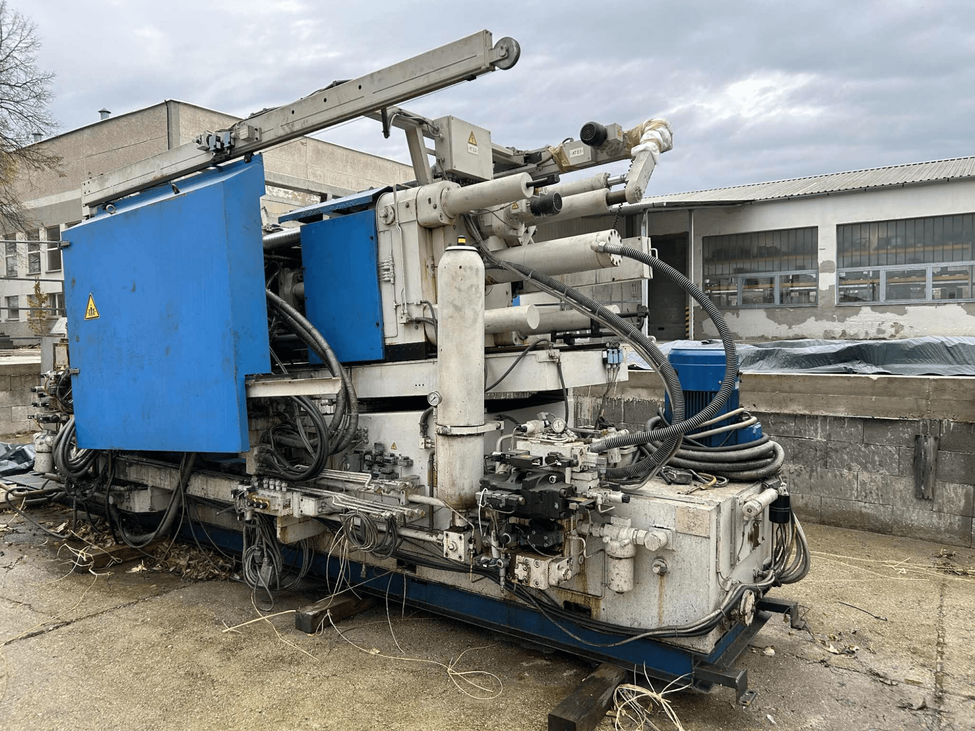 Didelė pramoninė hidraulinė mašina su mėlynu korpusu, įvairiomis žarnomis ir metaliniais komponentais, žiūrint iš šono.