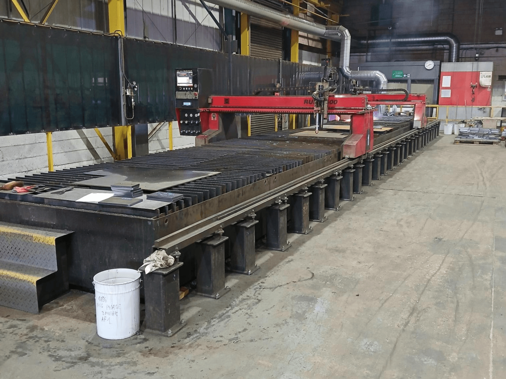 Raudonos spalvos pjovimo staklės RUIDA 300, vaizdas iš šono, su metalo lakštais ant plieninio stalo, pramoninėje darbo vietoje.