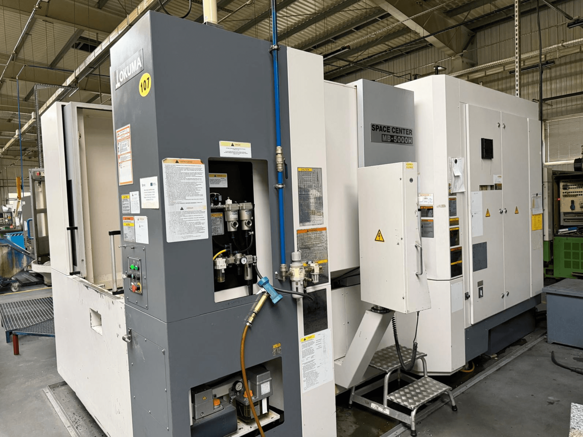 "Okuma MB 5000H" horizontalusis apdirbimo centras, šoninis vaizdas, kuriame matomas valdymo skydelis ir mašinų komponentai gamykloje.