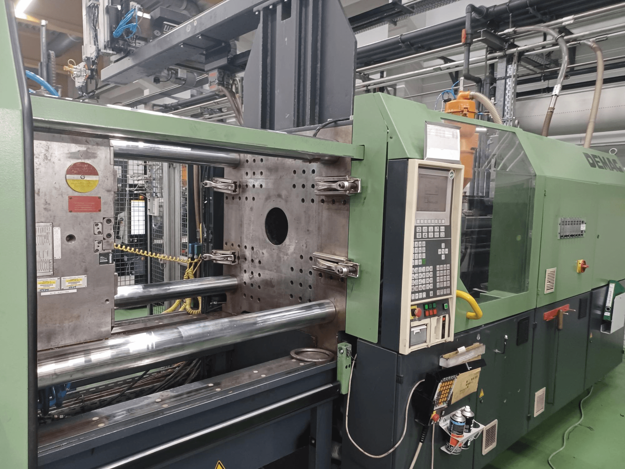 "Demag" įpurškimo liejimo mašina, vaizdas iš priekio, su valdymo pultu, metaliniais hidrauliniais cilindrais ir saugos funkcijomis.