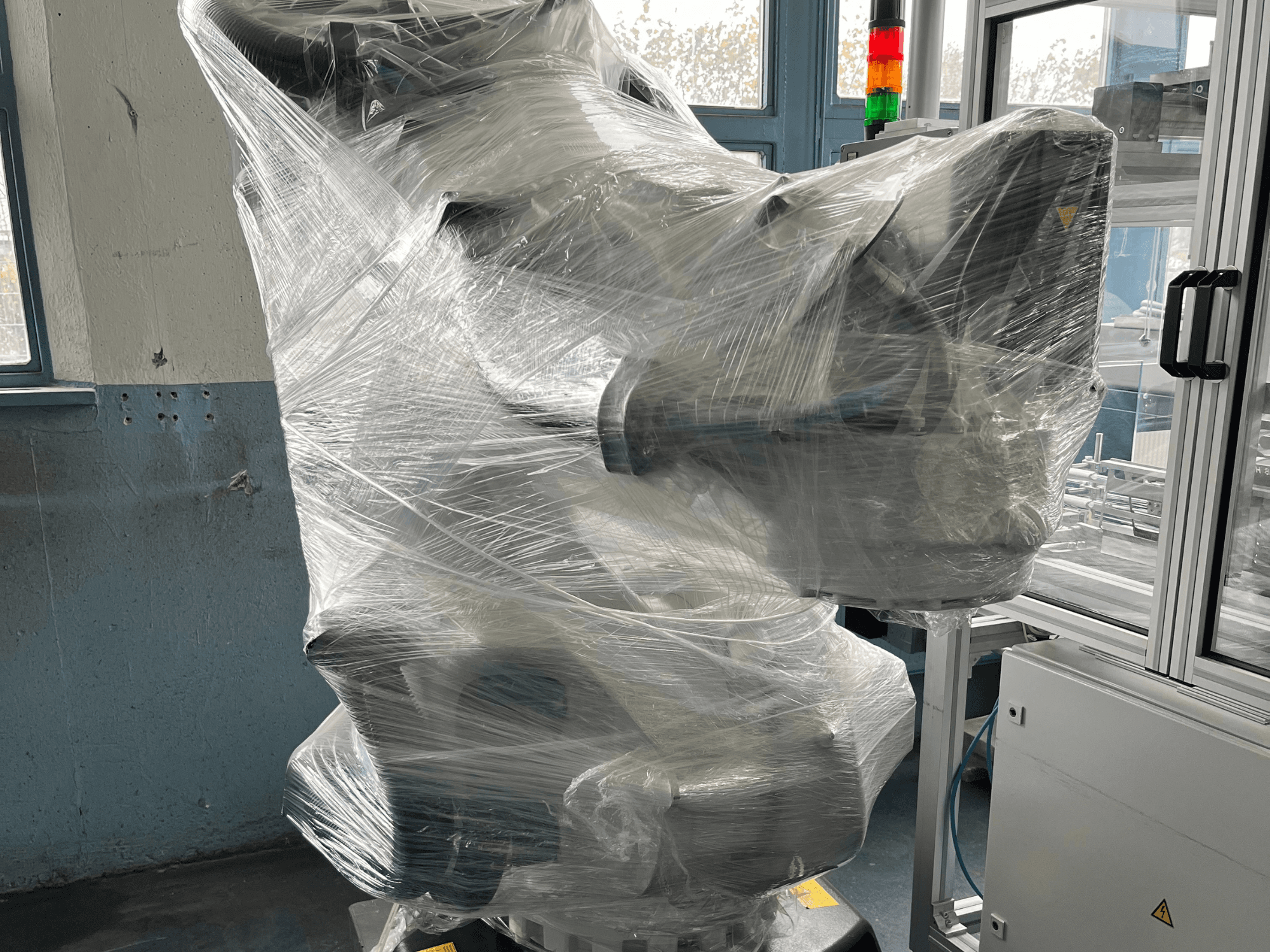 Pramoninė roboto ranka, apvyniota plastiku, vaizdas iš priekio. Į komplektą įeina valdymo blokas ir kontrolinė lemputė. Prekės ženklo nematyti.