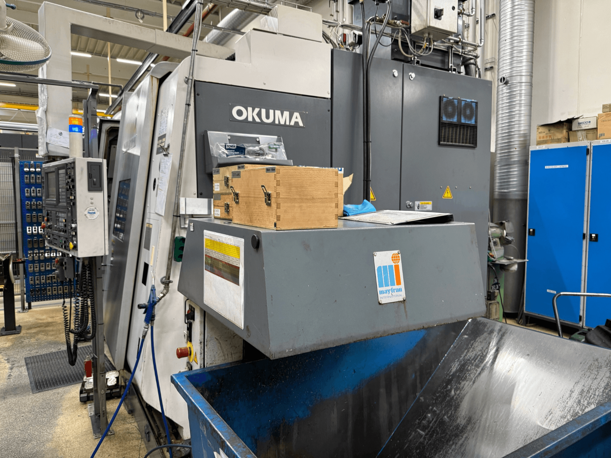 "Okuma" CNC staklės gamykloje, vaizdas iš priekio; valdymo skydelis, medinės dėžės ir mėlyna atliekų dėžė su "Mayfran" logotipu.