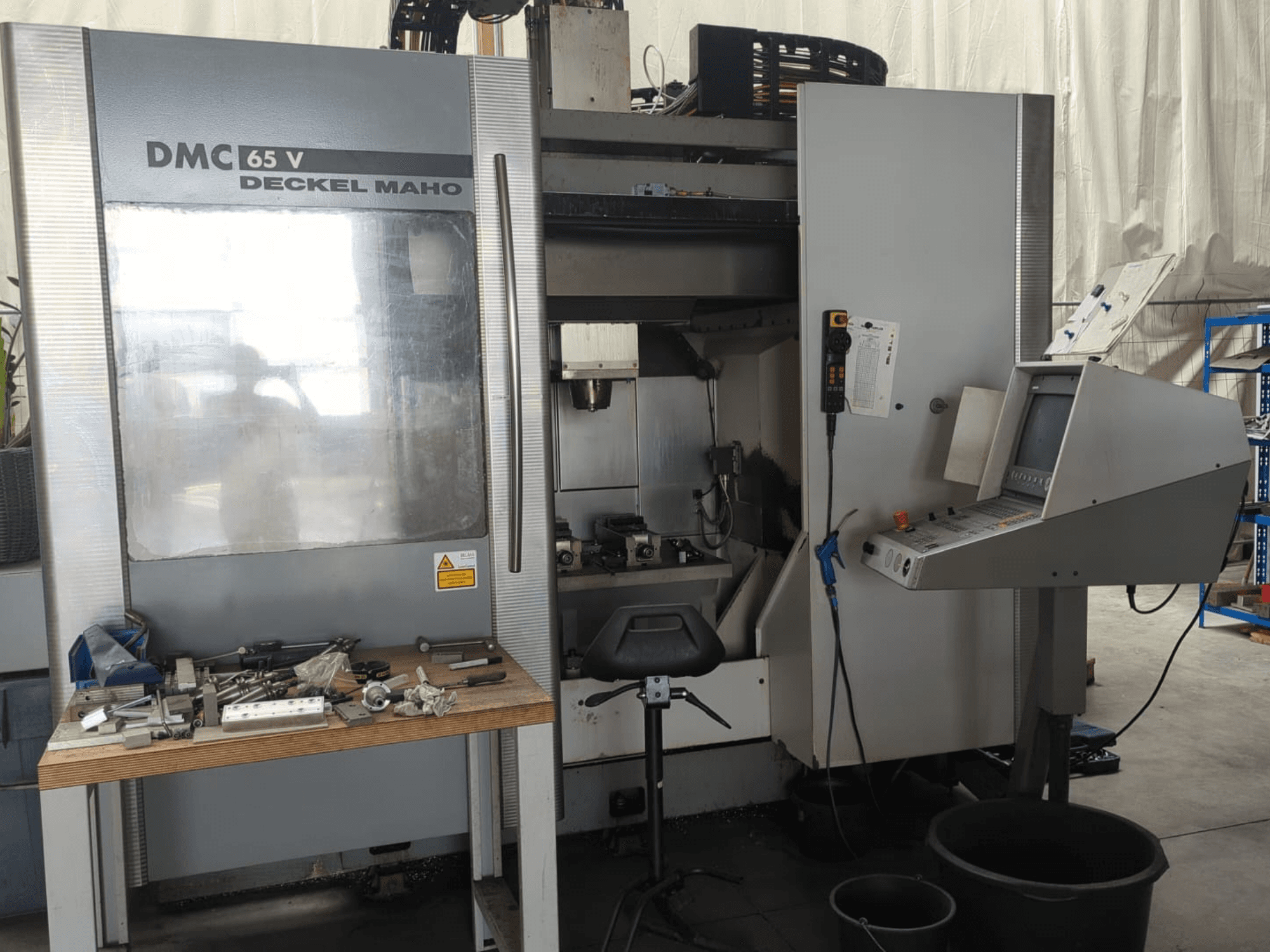 DECKEL MAHO DMC 65 V vertikaliojo apdirbimo centro vaizdas iš priekio su valdymo pultu, ant stalo esančiais įrankiais ir darbo vietos išdėstymu.