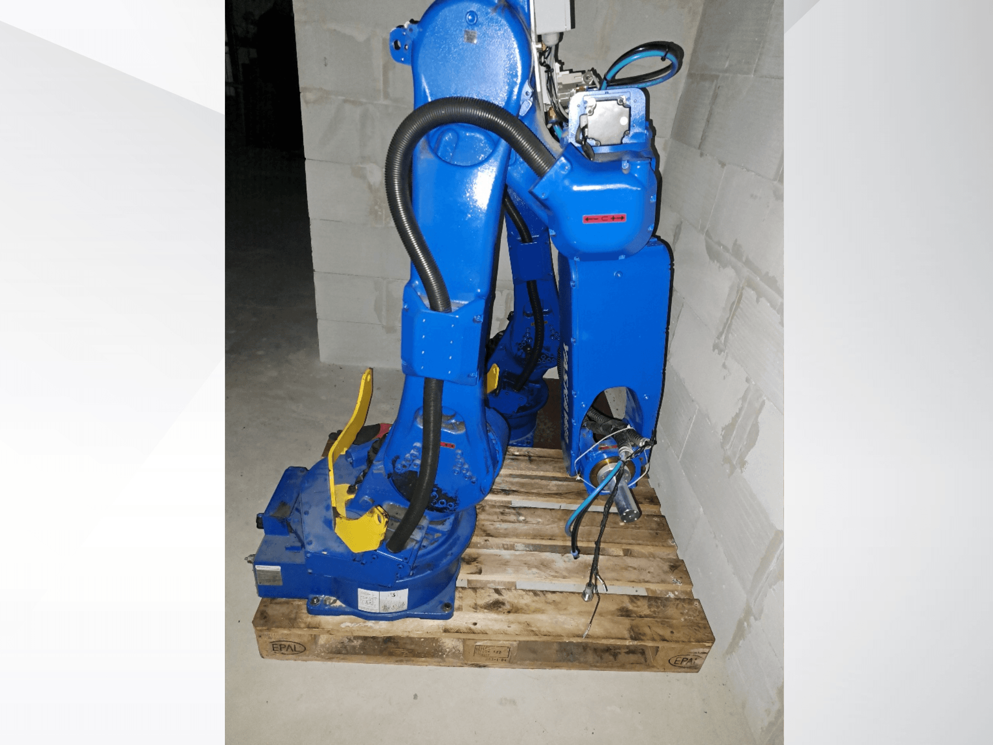 Mėlynos spalvos „Yaskawa Motoman GP25“ roboto ranka, vaizdas iš šono ant medinio padėklo, matomi kabeliai ir mechaniniai jungimai.