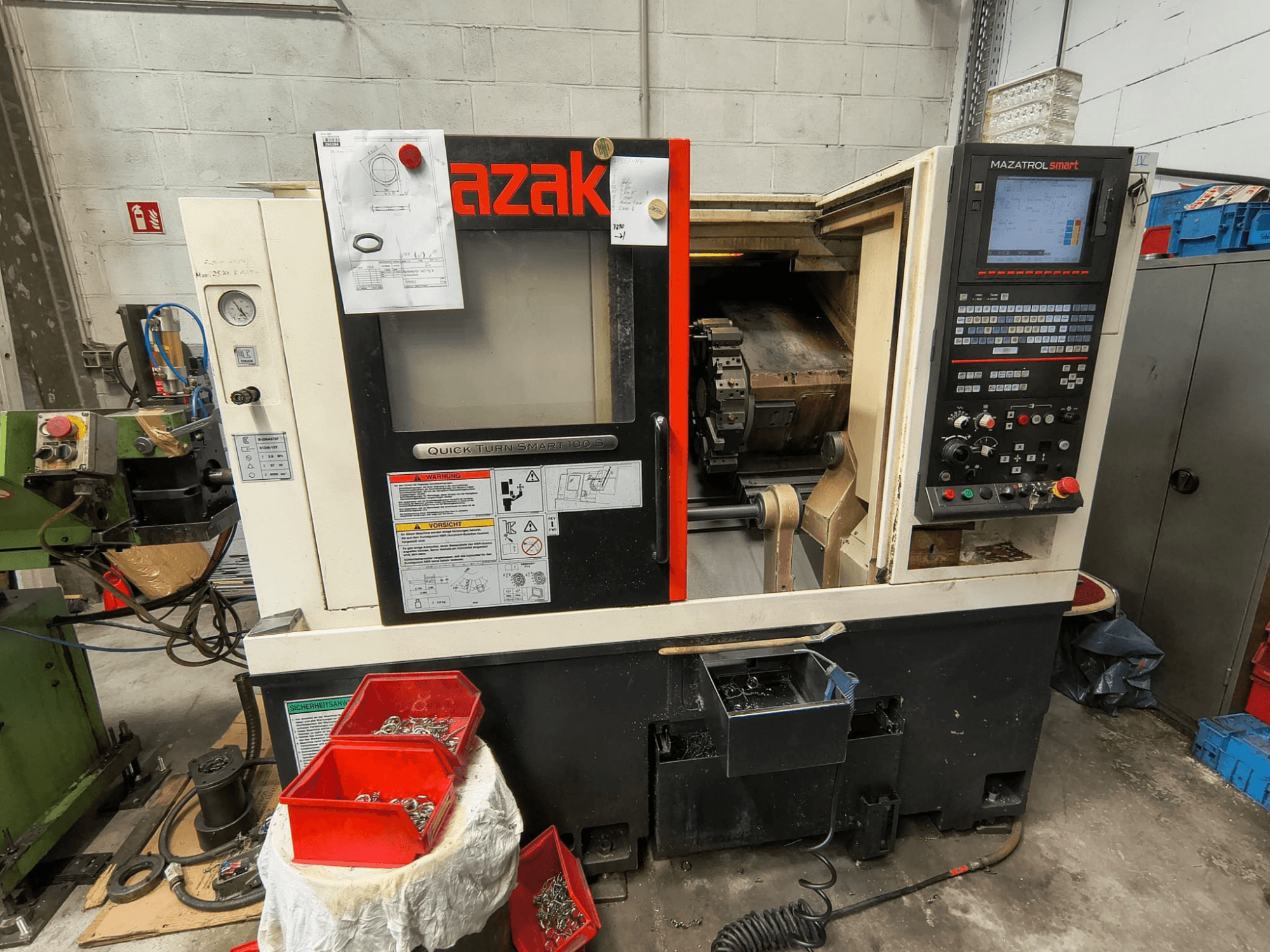 „Mazak Quick Turn Smart 100 S“ – šoninis vaizdas, kuriame matomas valdymo skydelis, aušinimo skysčio surinkimo dėklas ir laikymo dėžės su varžtais.