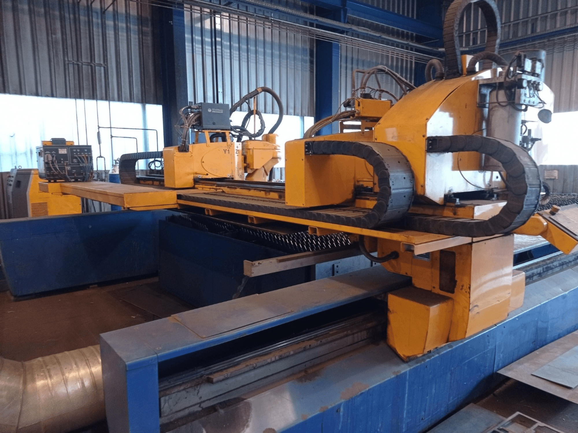 Geltonos spalvos CNC plazminio pjovimo staklės su dviem horizontaliomis rankomis, parodytos iš priekio, su valdymo pultu ir pjovimo lova.