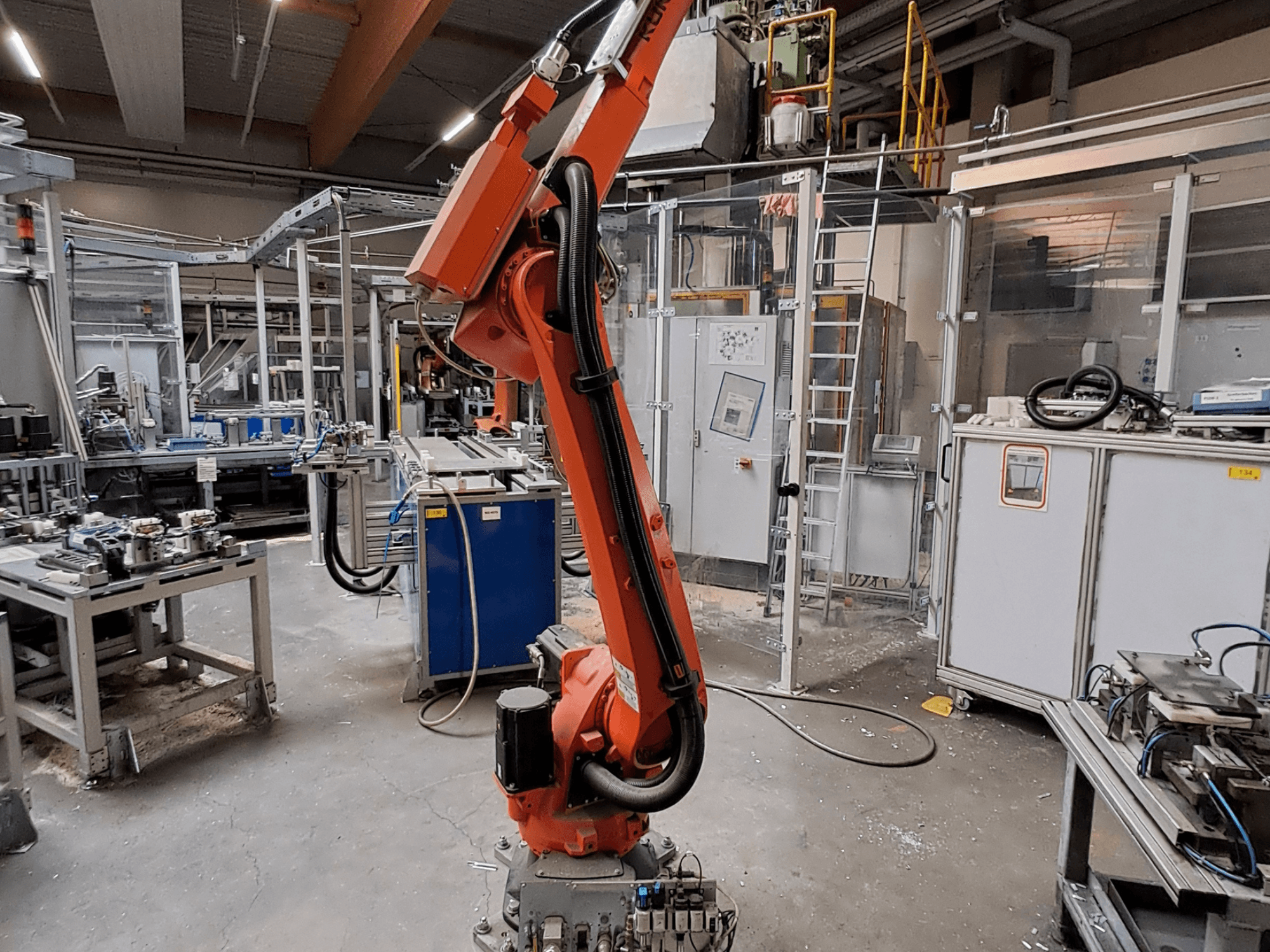 Pramoninėje aplinkoje esanti „KUKA KR 26 R3100/SEL“ robotinė ranka, pavaizduota iš šono, o fone matoma darbo įranga.