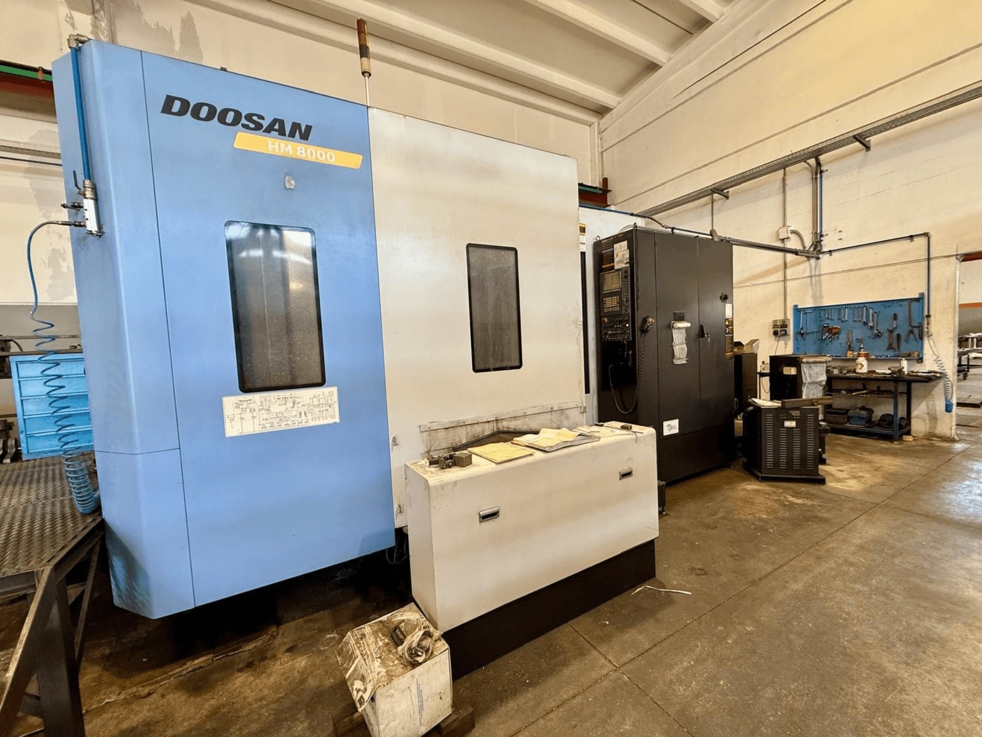 Mėlynos spalvos „DOOSAN HM 8000“ horizontalus apdirbimo centras, vaizdas iš priekio, kuriame matomas valdymo pultas ir šalia esanti dirbtuvės įranga.