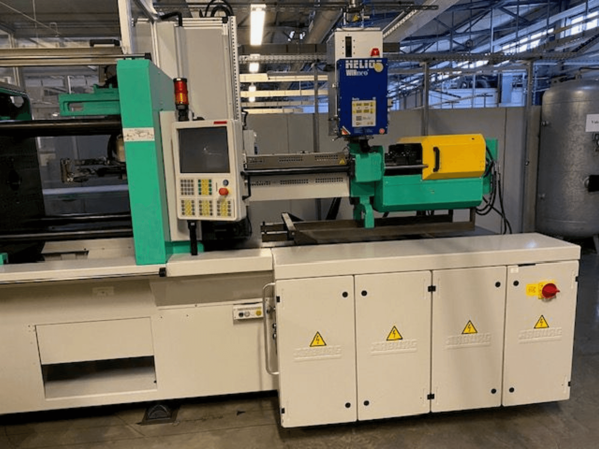 HELIO modelio pramoninė liejimo mašina, vaizdas iš priekio, su valdymo skydeliu, apsauginiais skydais ir avarinio stabdymo mygtuku.