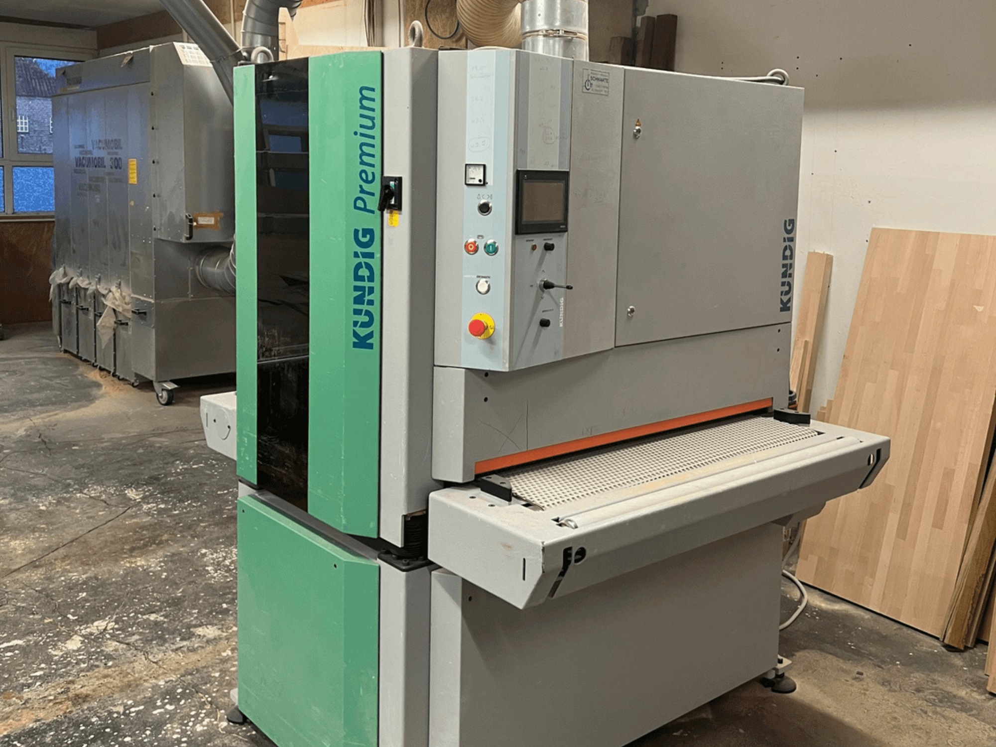 Pilkos ir žalios spalvos juostinis šlifuoklis "Kundig Premium-1 1100K", parodytas iš šono, matomas valdymo skydelis ir šlifavimo juosta.