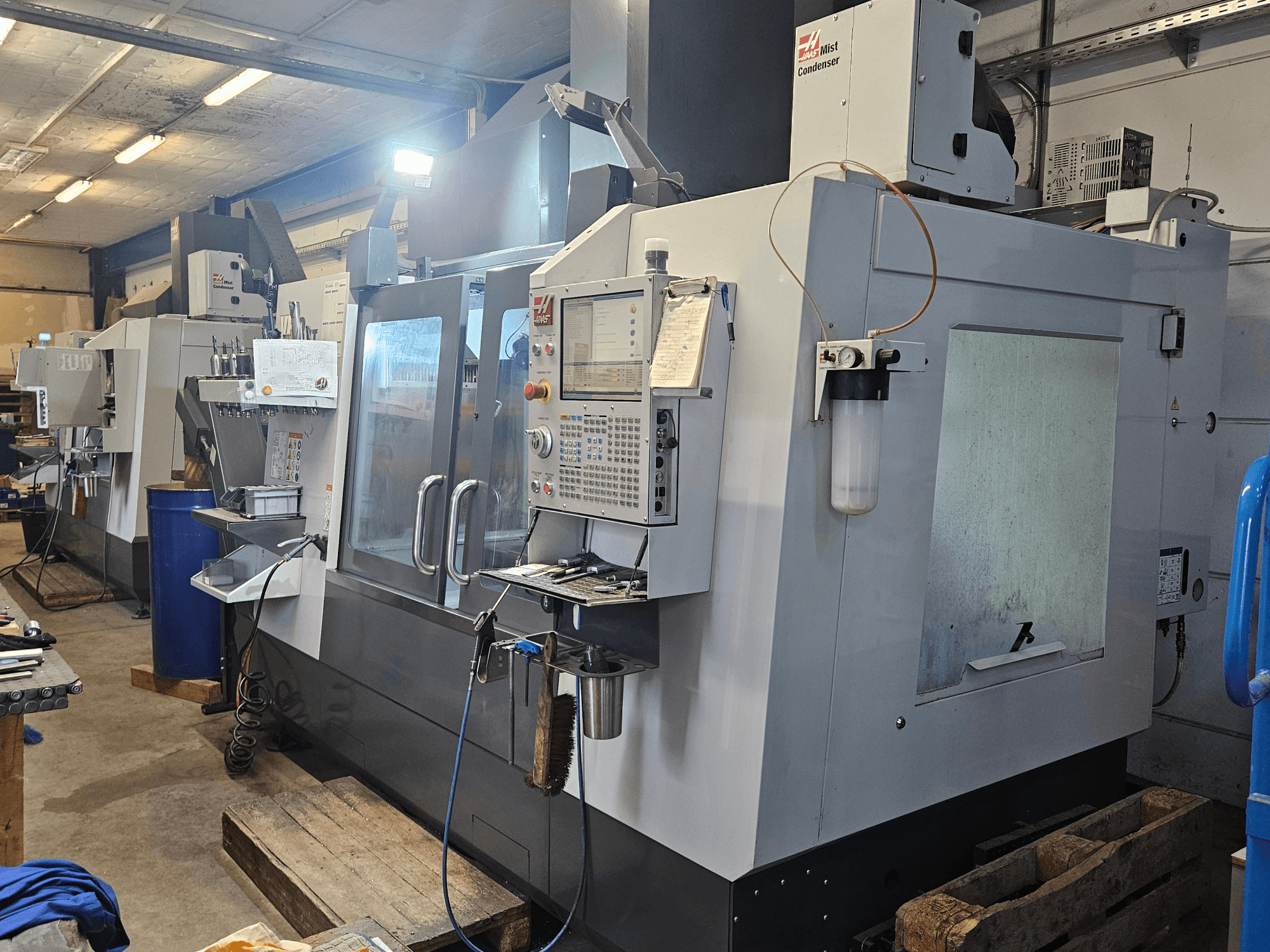 Vertikalus apdirbimo centras „Haas VF-3YT/50“ dirbtuvėse, vaizdas iš priekio, kuriame matomas valdymo pultas, įrankių laikikliai ir garų kondensatorius.